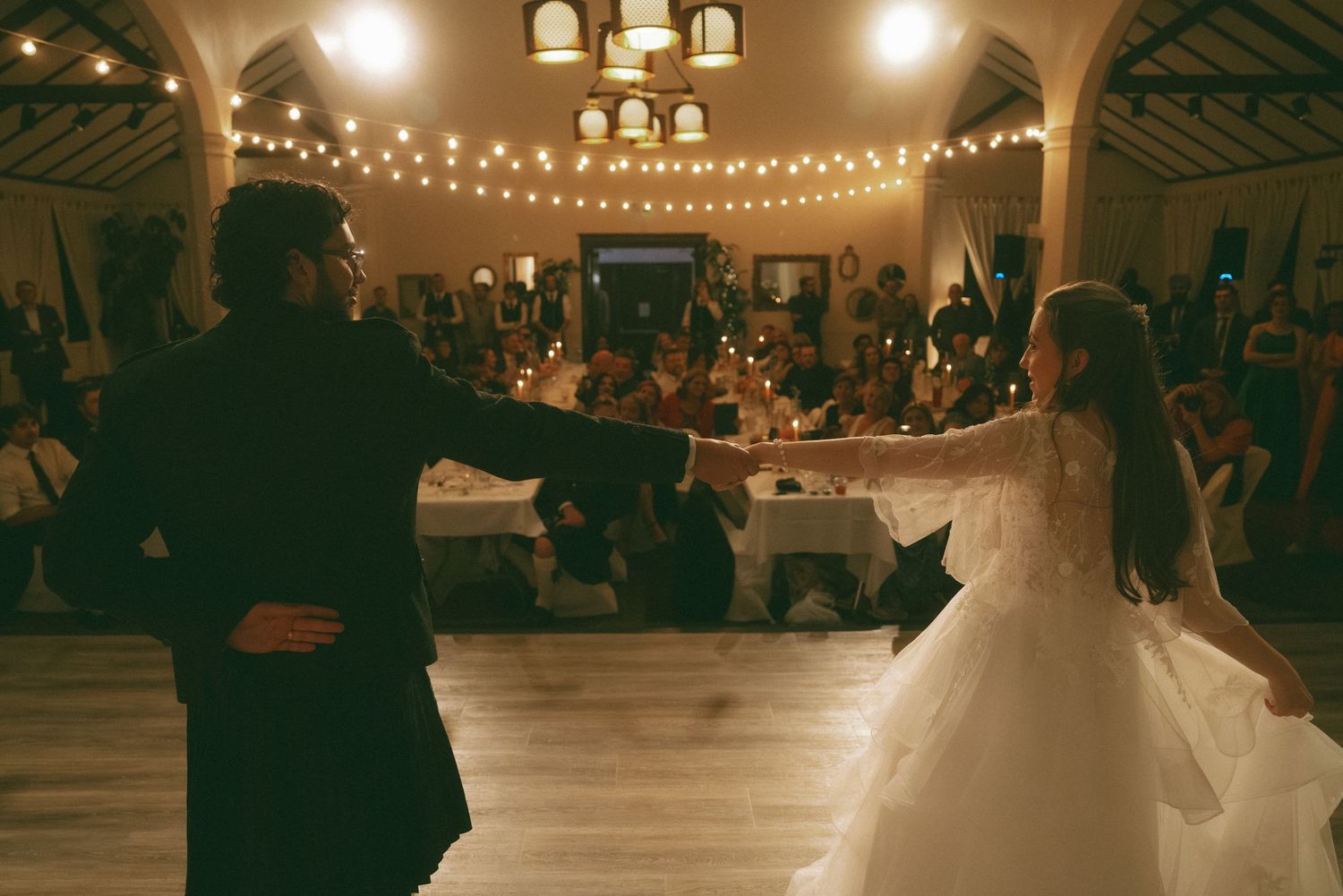 Wedding reception dance under string lights in an elegant venue with arched ceilings.