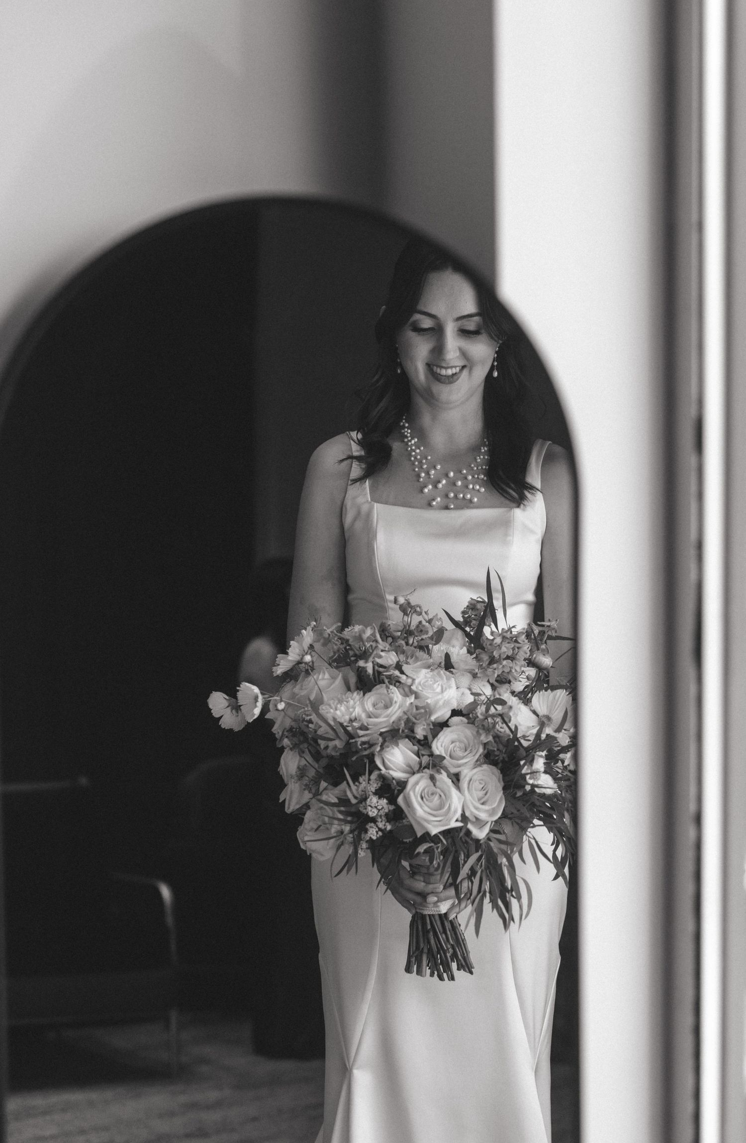 A bride in a sleek white dress holds an elegant bouquet of roses and mixed flowers.