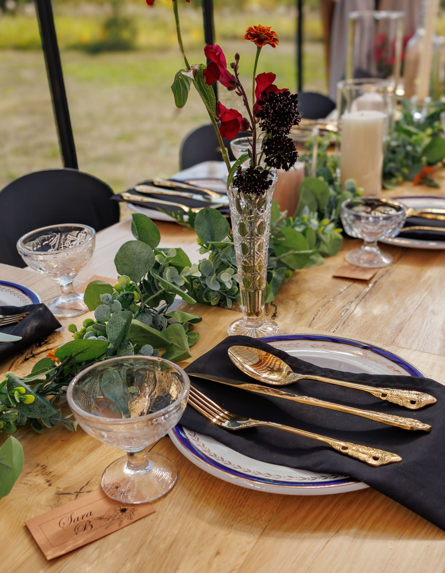 Rustic wedding table decor with black napkins, gold cutlery, and small floral centerpieces on wooden tables.