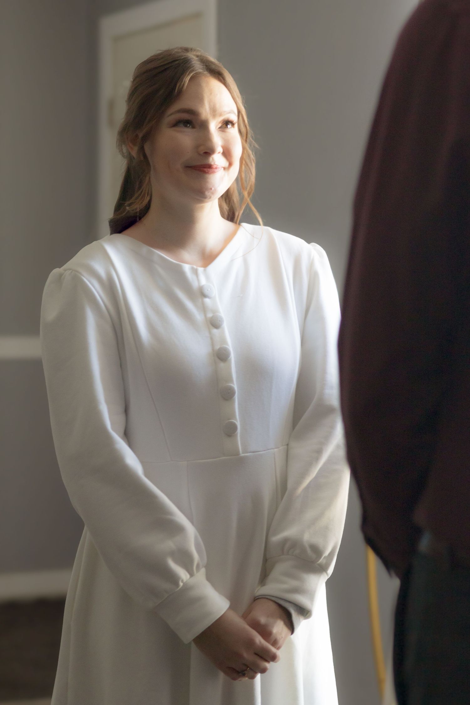 A person in a white dress stands indoors near natural light.