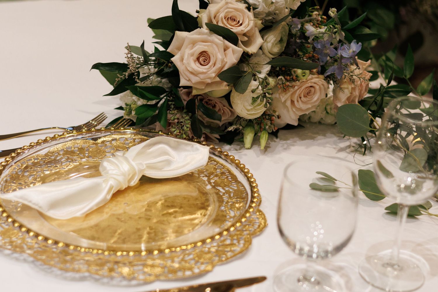 Elegant table settings with gold charger plates, blush roses, and greenery create a romantic wedding reception atmosphere.