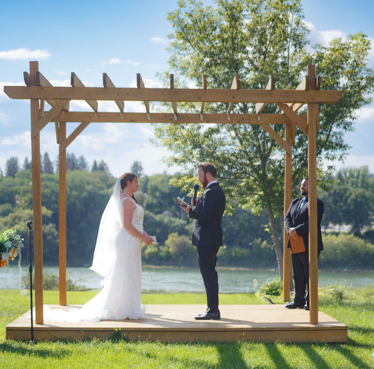 Wedding ceremony under wooden pergola with lake and trees in background.