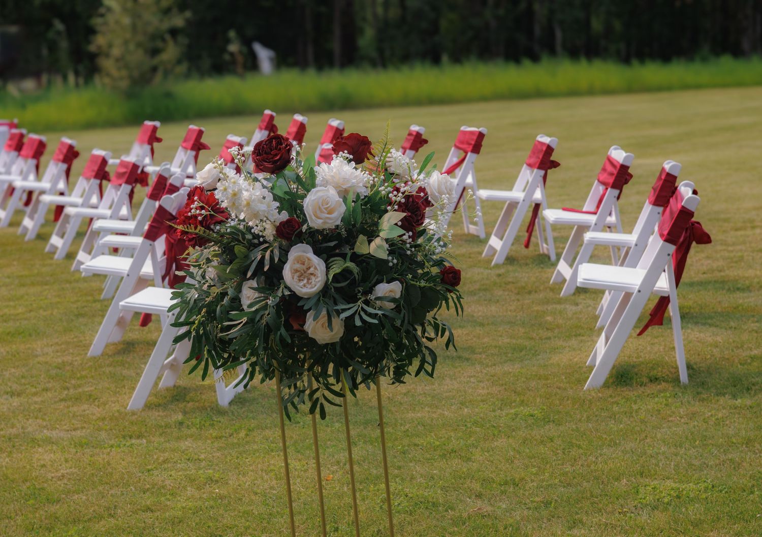 White wedding chairs decorated with red flowers and greenery set up for an outdoor ceremony.