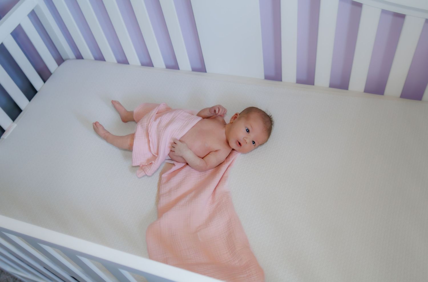Infant in pink sleepwear resting peacefully in a purple and white crib.