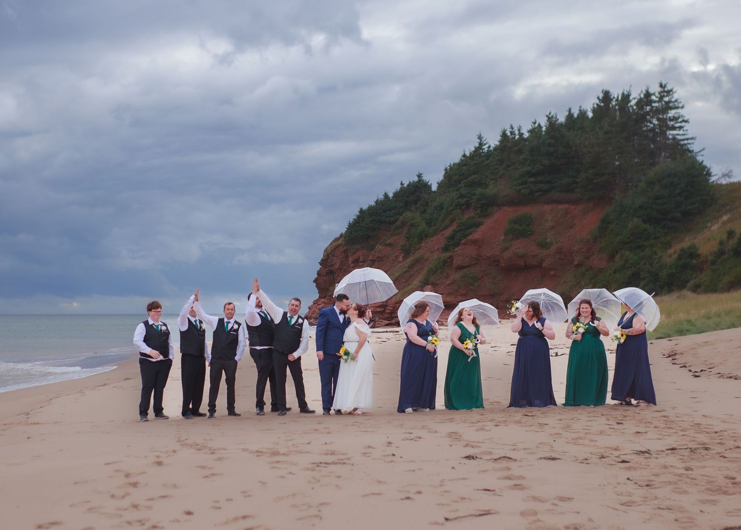 Wedding party takes photos on a beach with red cliffs and ocean in the background.