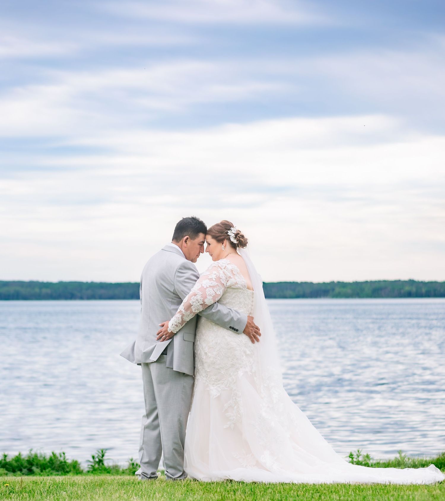 A romantic wedding couple embraces on a grassy hill overlooking a serene lake on their special day.