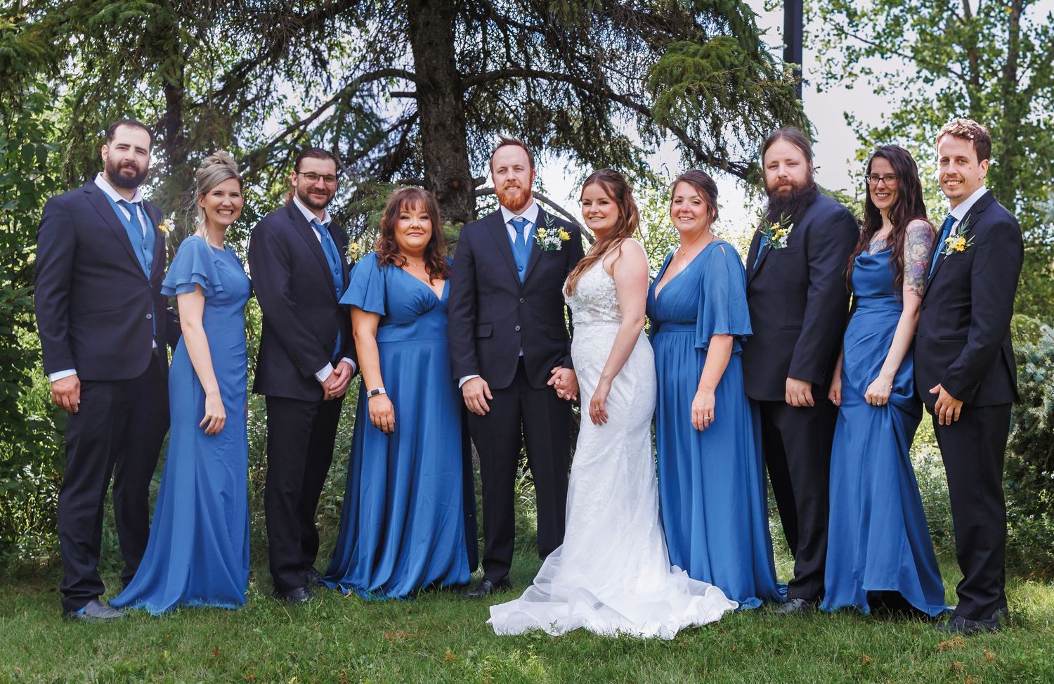 Wedding party in coordinated blue dresses and dark suits posed outdoors under trees.