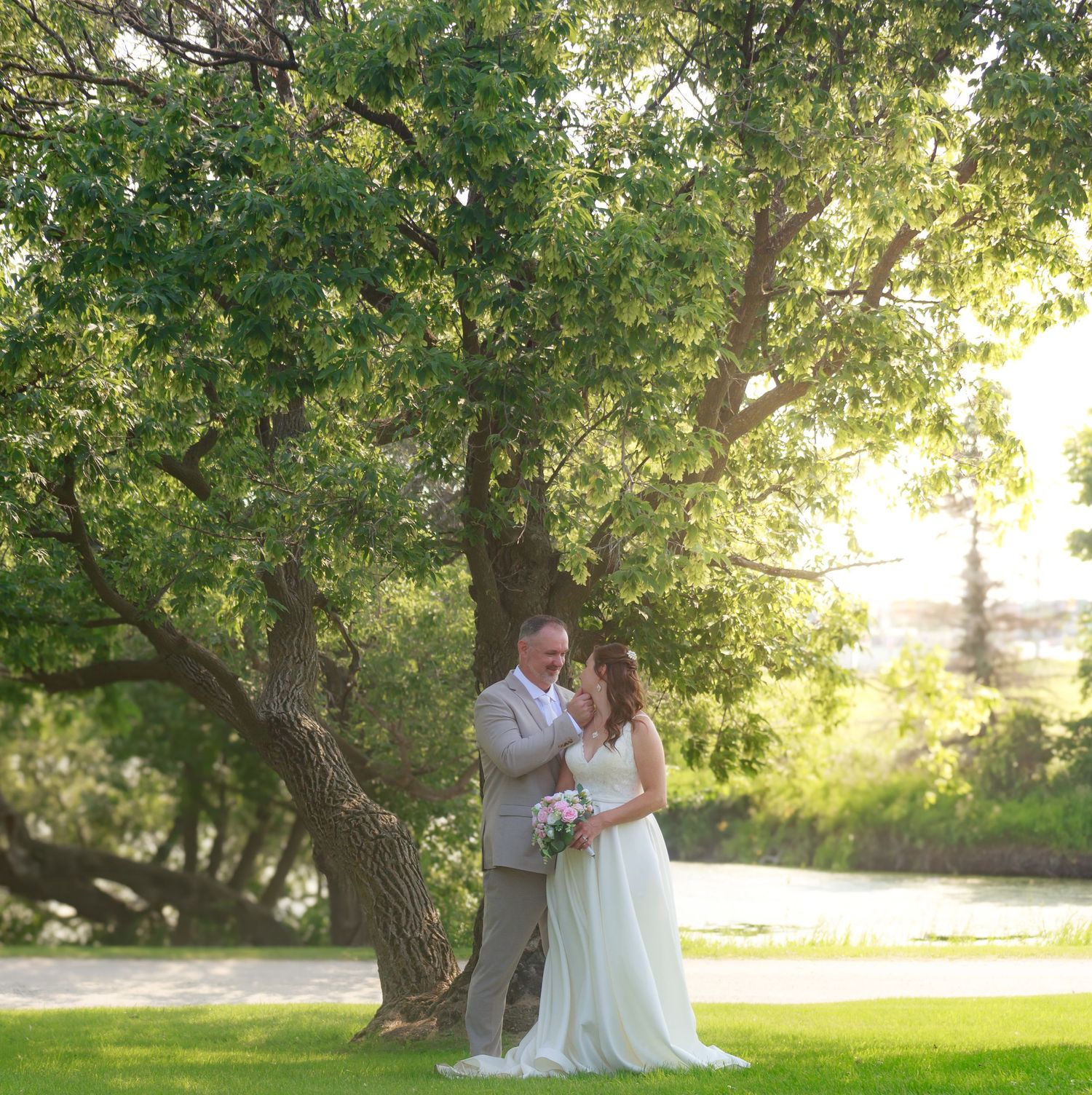 A couple shares an intimate moment under green trees in a romantic outdoor wedding setting.