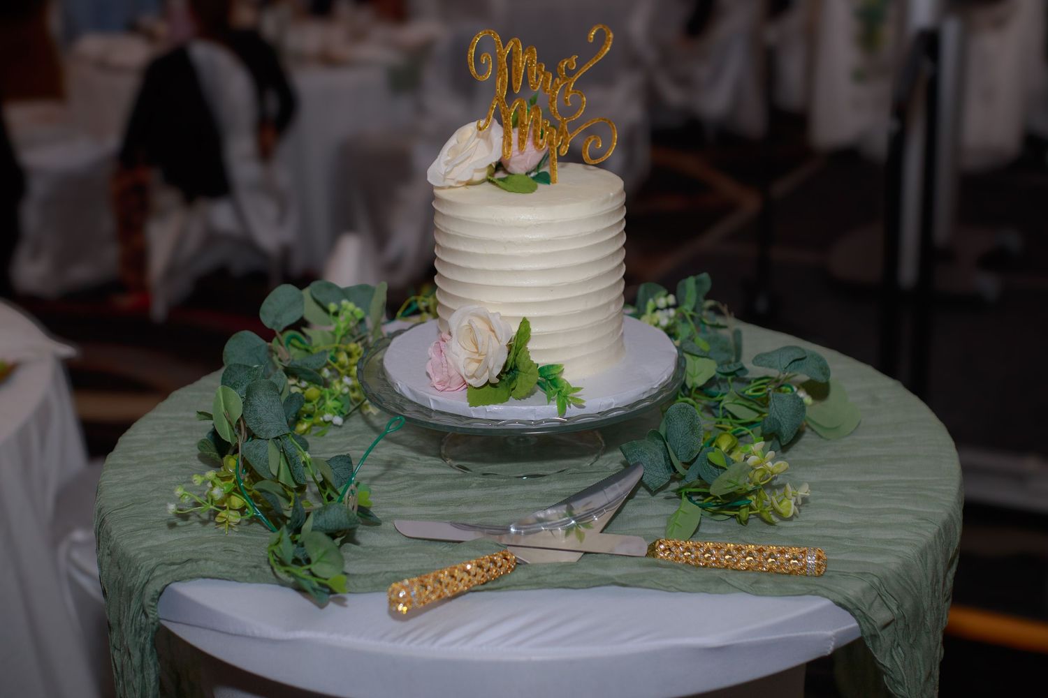White wedding cake decorated with greenery and gold accents on a mint green tablecloth.