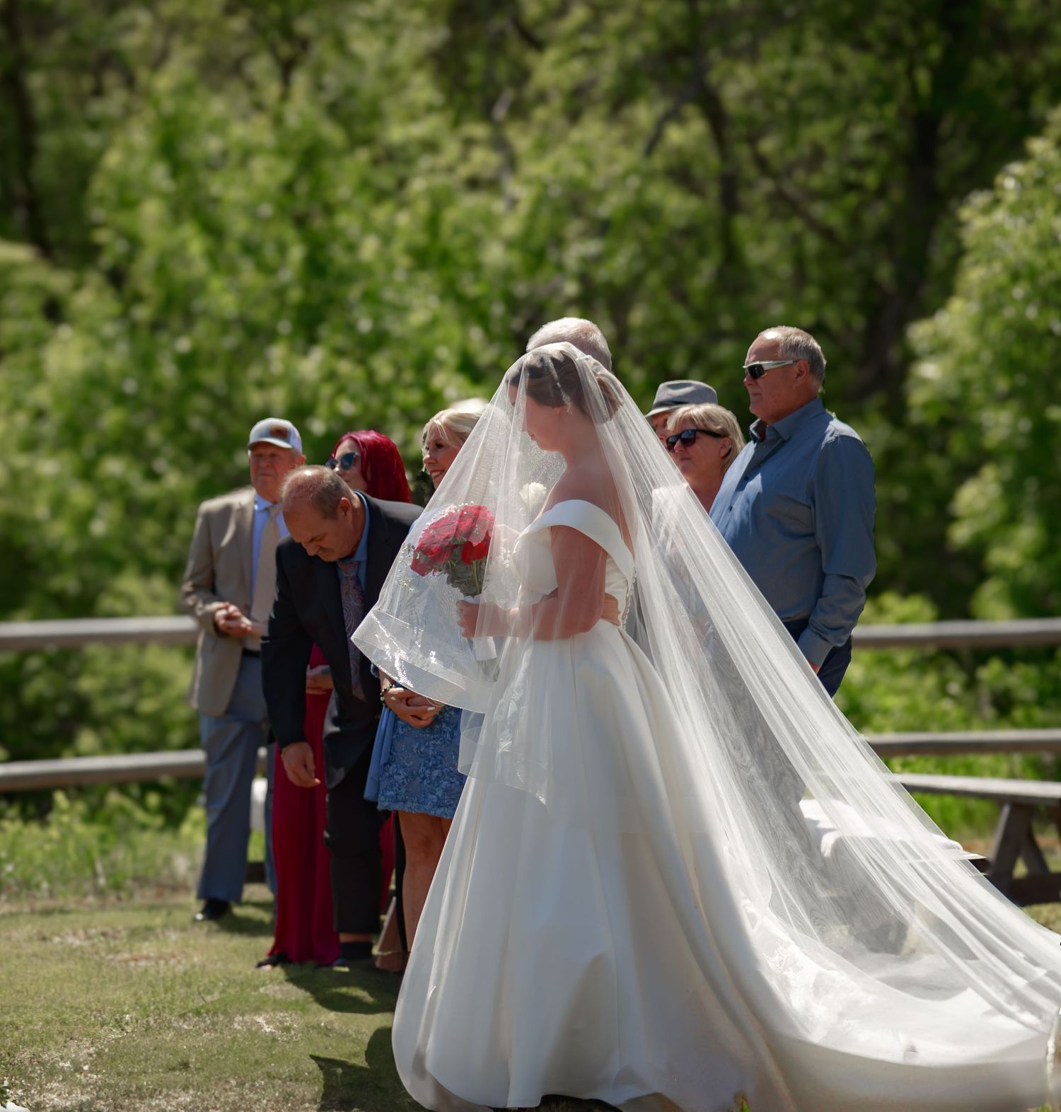 Wedding party gathers outdoors under trees with flowing white bridal gown and veil.