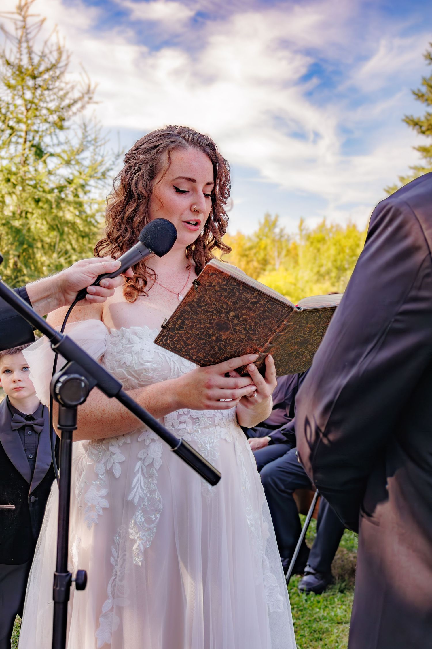 Wedding ceremony moment captured outdoors with guests under blue sky and evergreen trees.