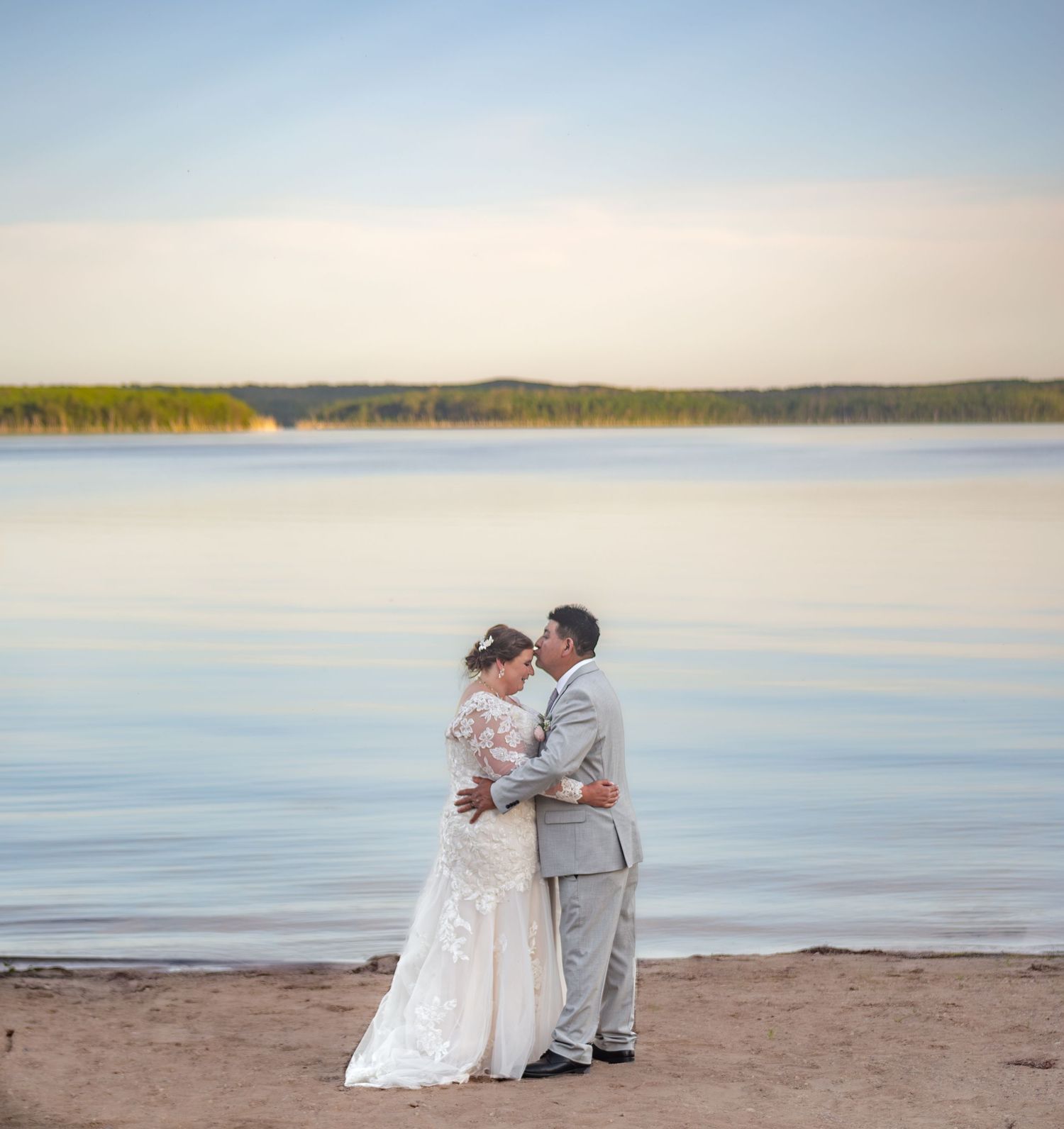 A wedding couple embraces on a sandy beach with calm waters stretching to the horizon.