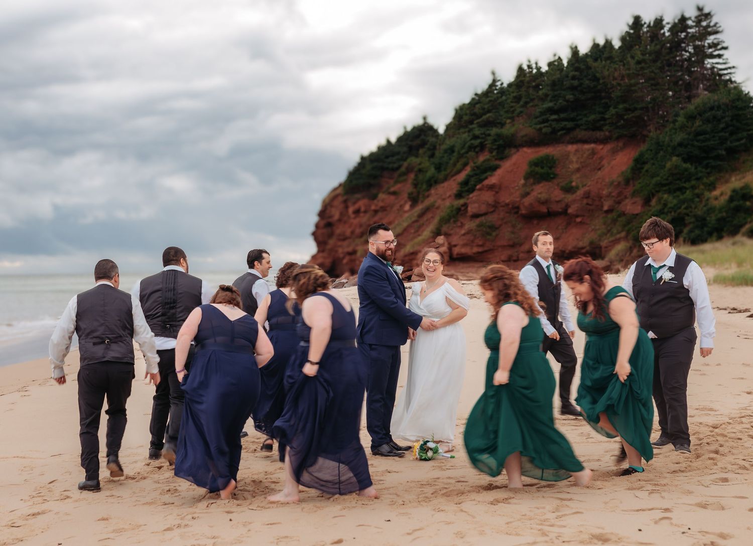 Wedding party sits casually on beach with red cliffs and ocean views.