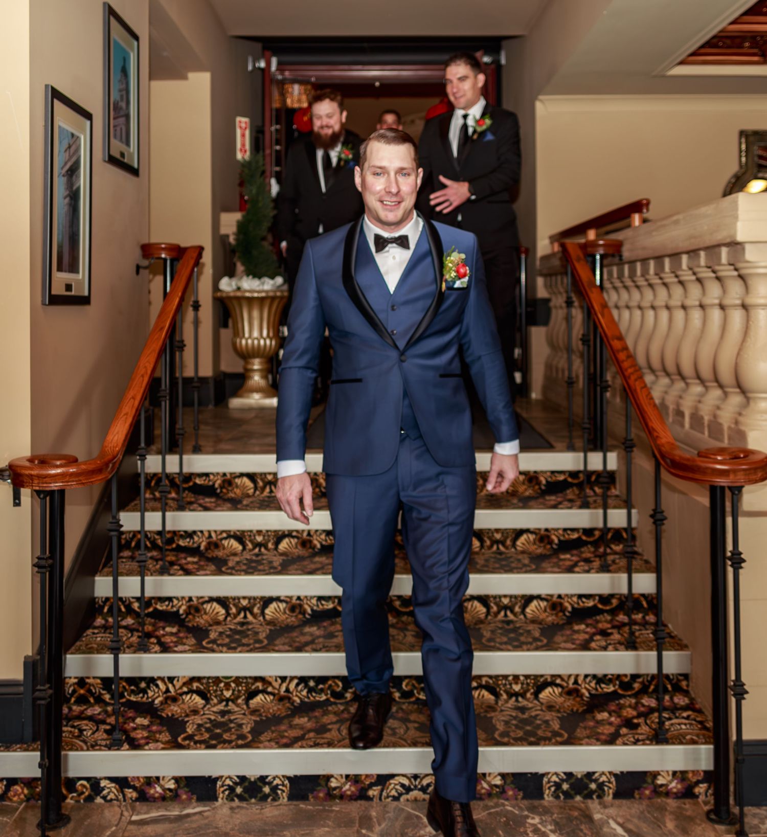 Groomsmen in navy suits walking down elegant carpeted stairs in formal venue.