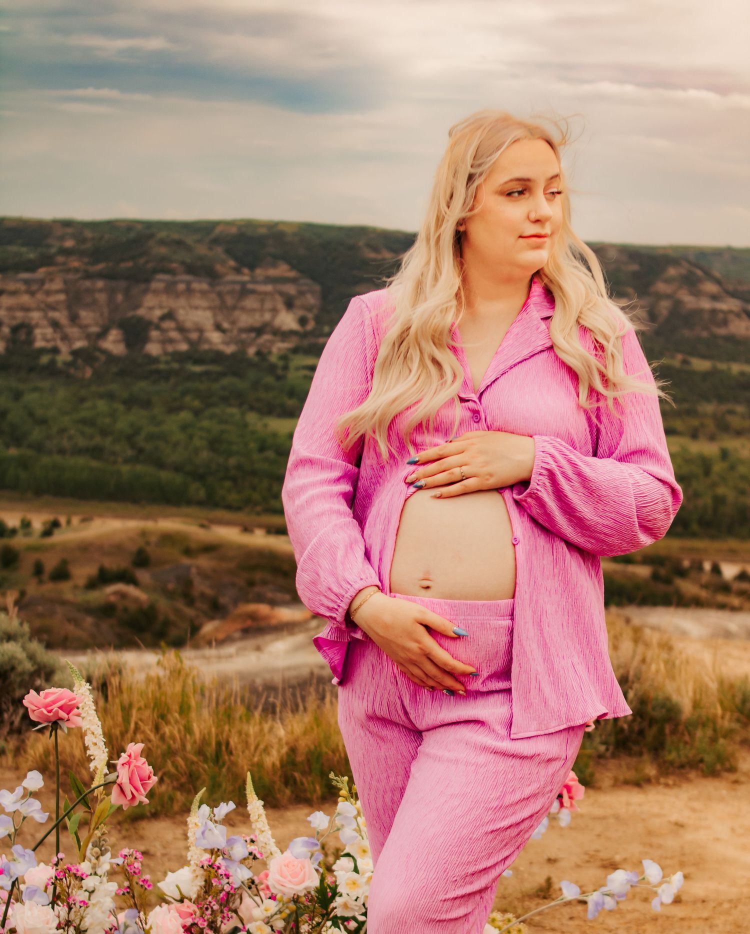 Maternity photoshoot featuring pink outfit against scenic mountain backdrop with floral decorations.