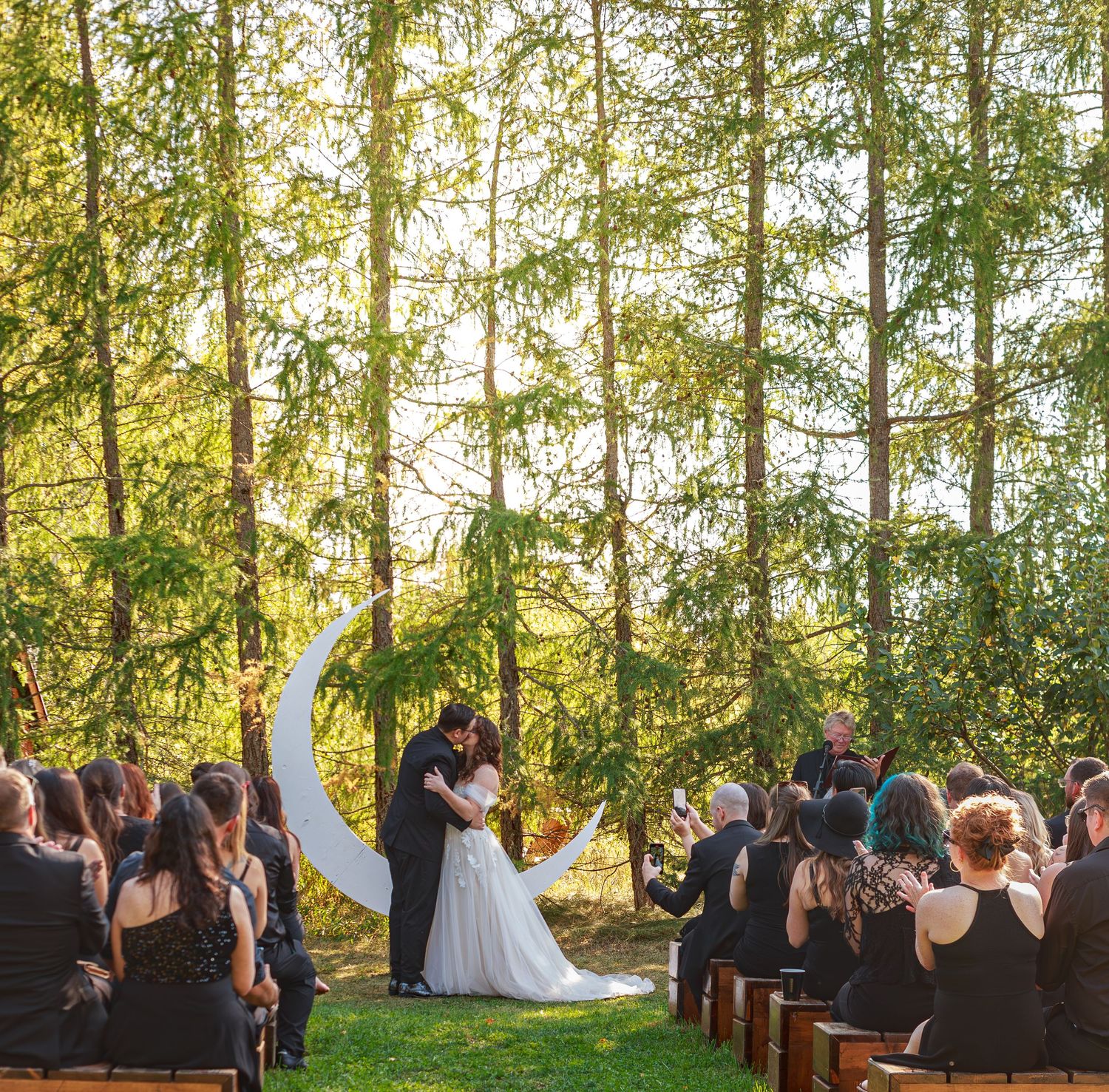 Wedding ceremony taking place in a serene forest setting with wooden benches and natural sunlight filtering through trees.