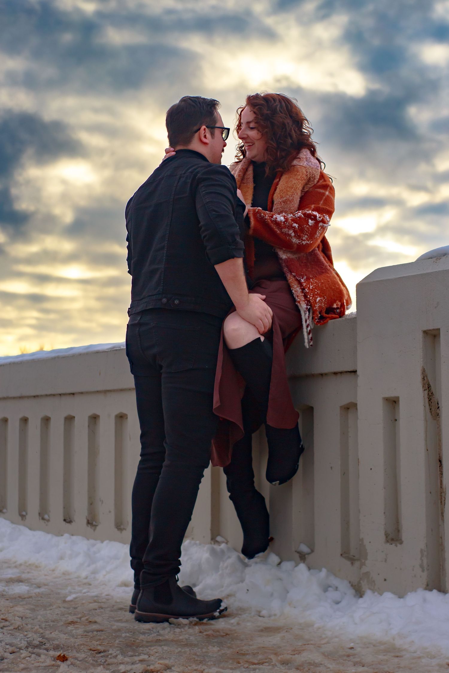 Couple embracing on snowy walkway with dramatic sunset sky in background.