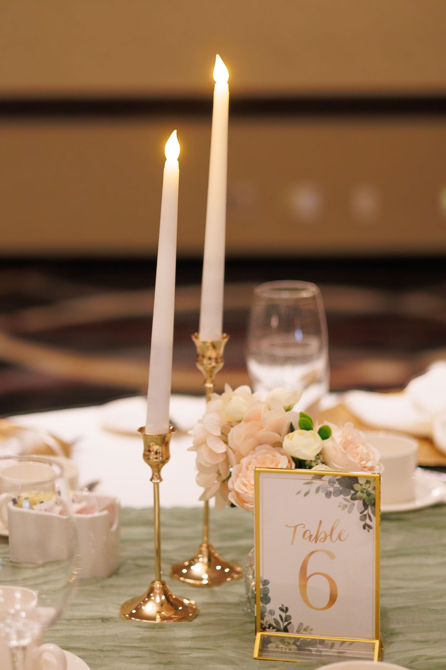 Elegant table setting with gold candlesticks, white flowers, and table number 6 in a gold frame.