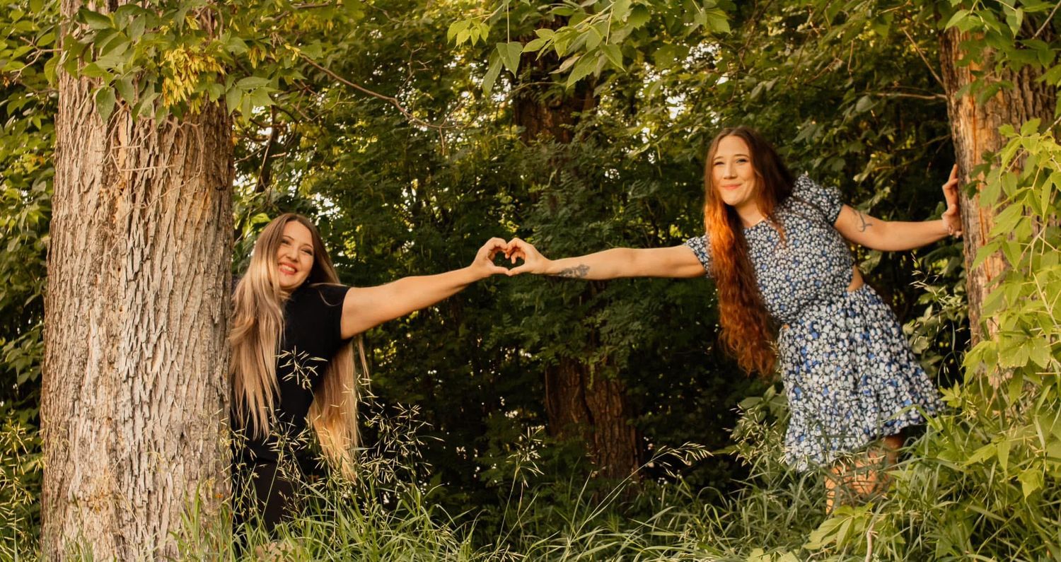 Two people making heart shapes with their hands between trees in a playful outdoor photo.