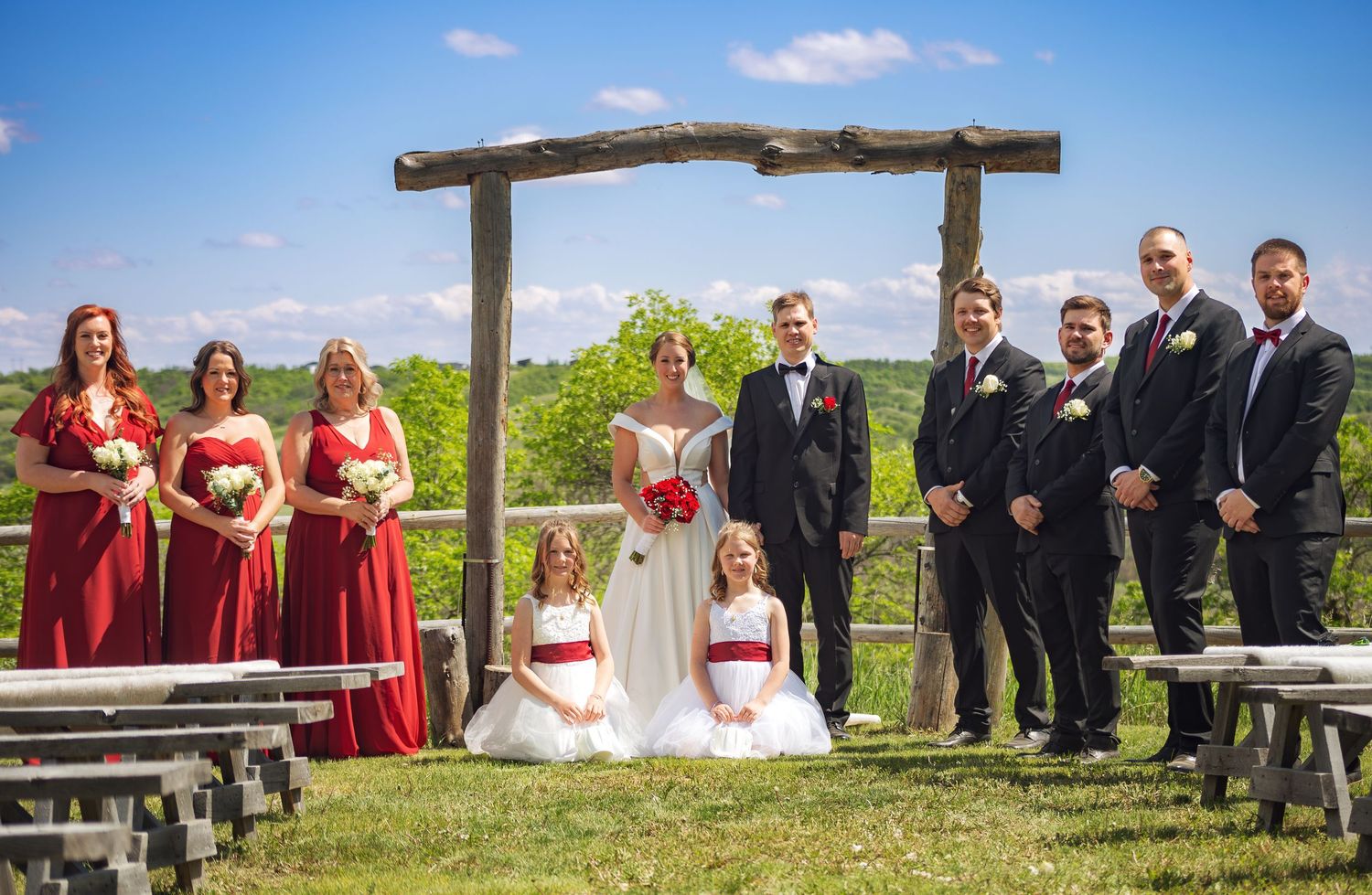 Outdoor wedding ceremony setup with wooden benches and rustic arch against blue sky.
