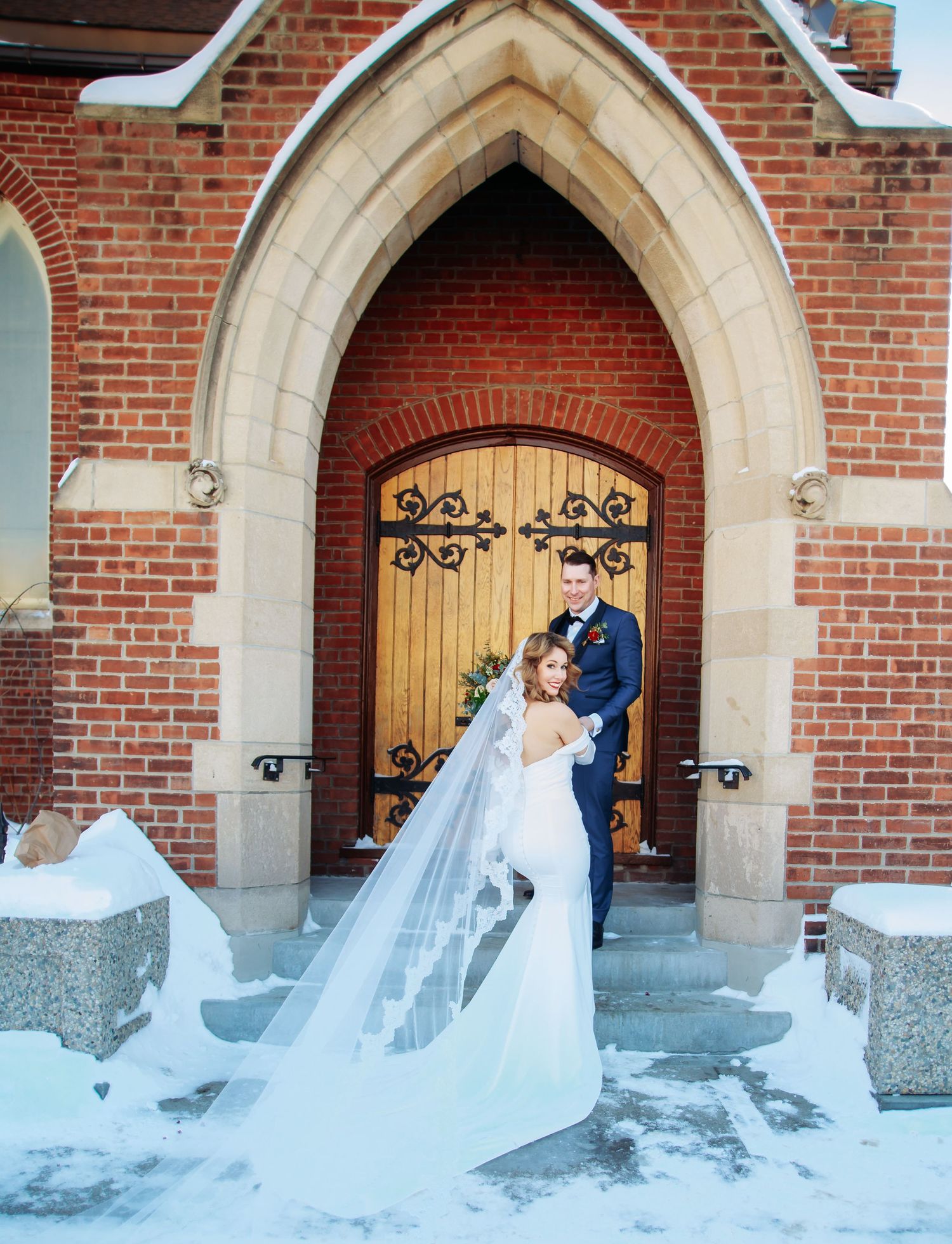 A winter wedding scene in front of a Gothic-style brick church with an ornate wooden door and stone archway.