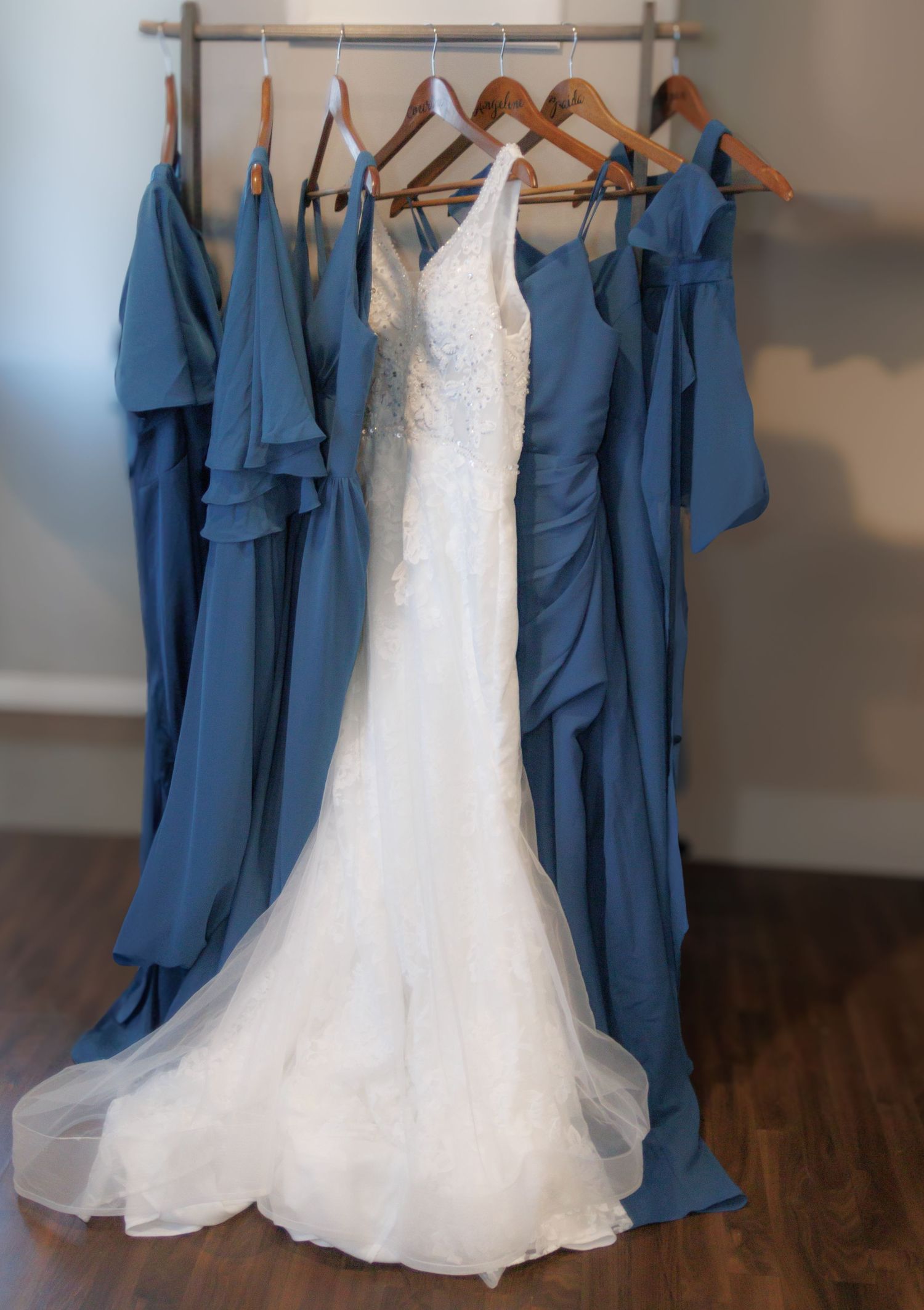 White wedding dress displayed among blue bridesmaid dresses on wooden hangers.