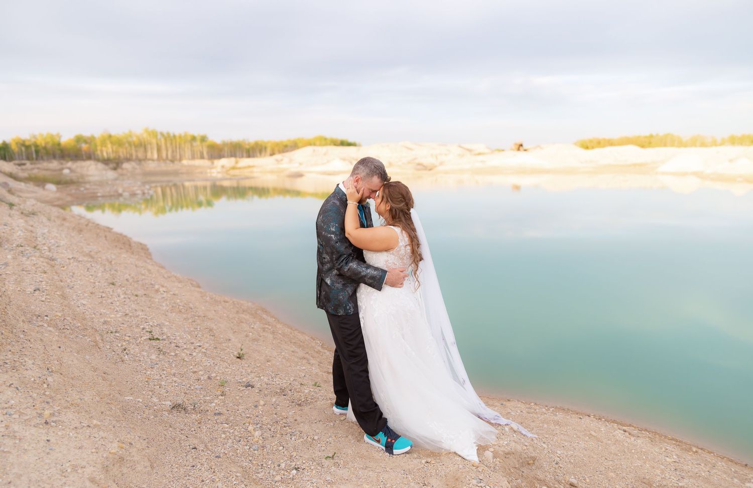 Wedding couple poses on sandy beach with turquoise water in background during golden hour.