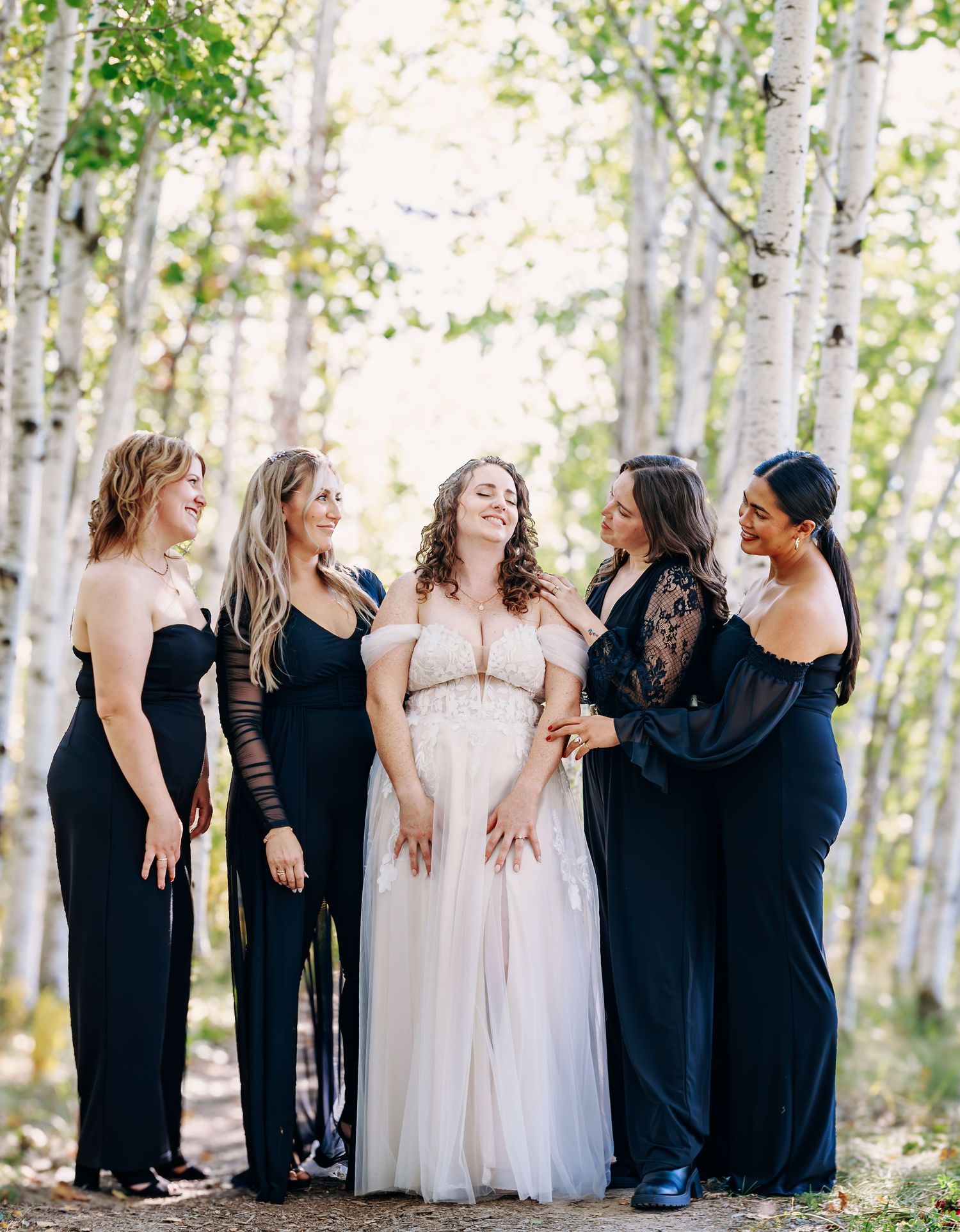 Bridesmaids in black dresses standing with bride in white gown among birch trees.