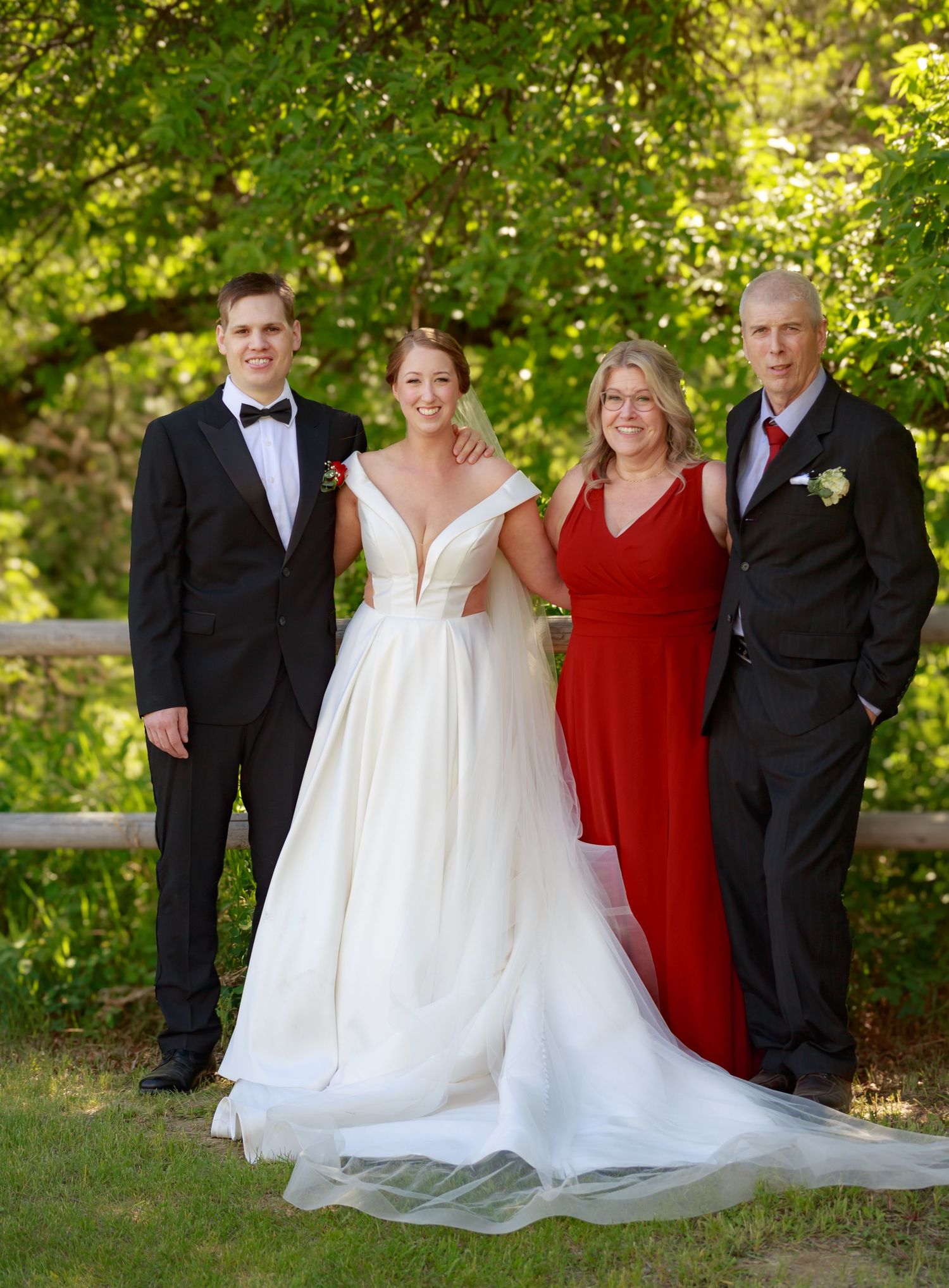 Wedding party in white gowns and black tuxedos posed outdoors with greenery.