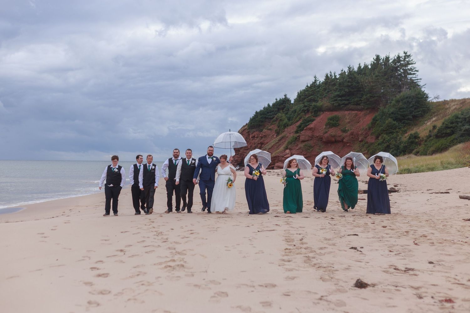 Wedding party walking along a beach shoreline under cloudy skies.