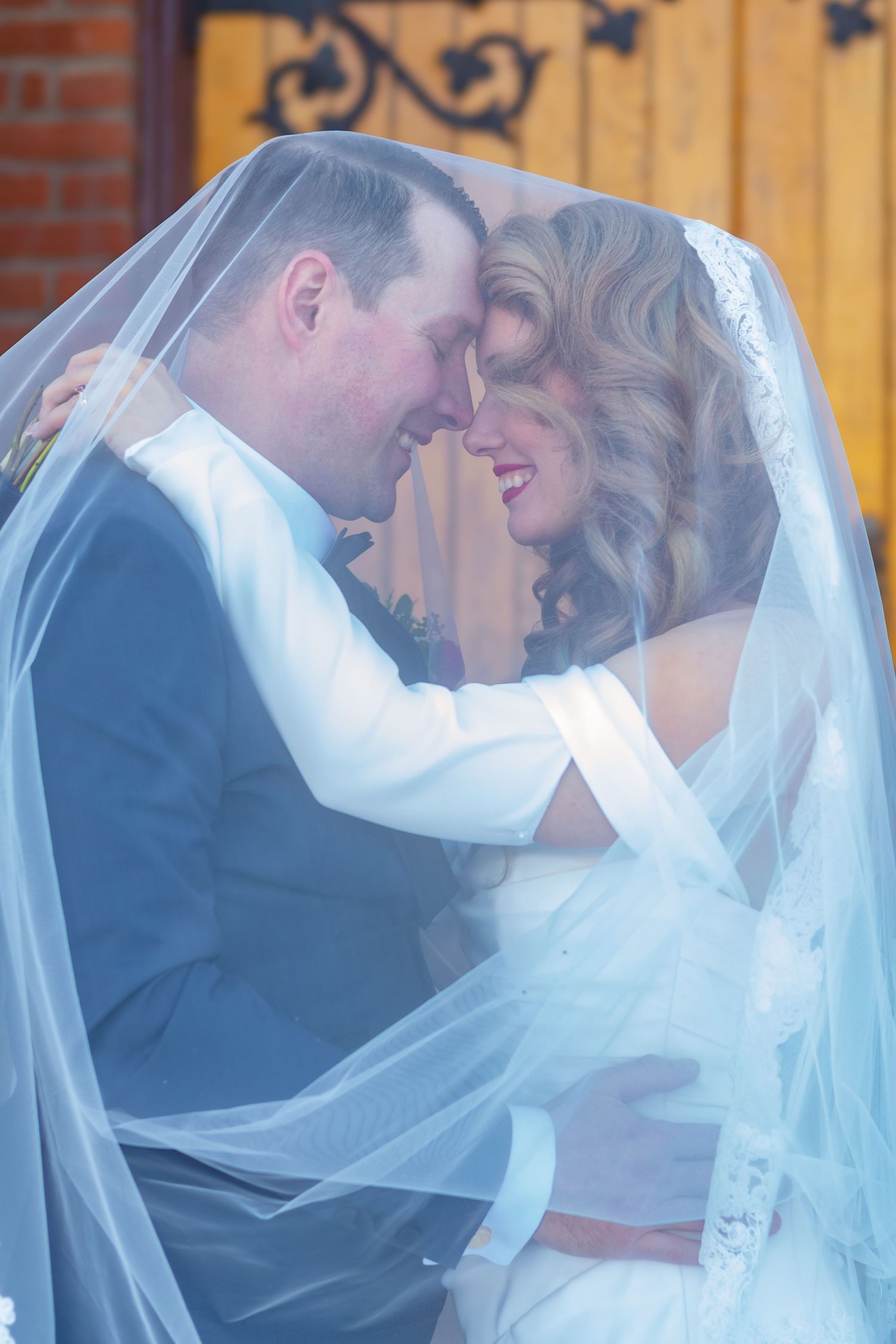 Romantic wedding moment captured through flowing white veil against brick architecture.