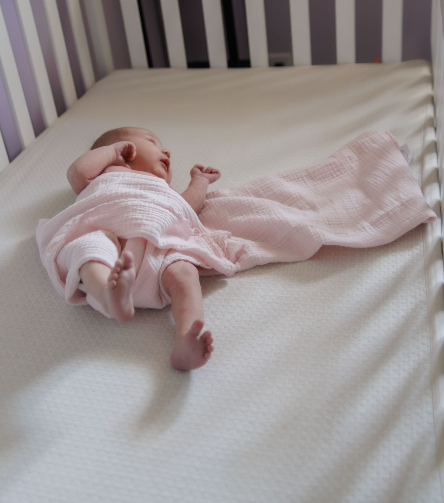 Baby sleeping soundly on white bedding in a purple-barred crib.