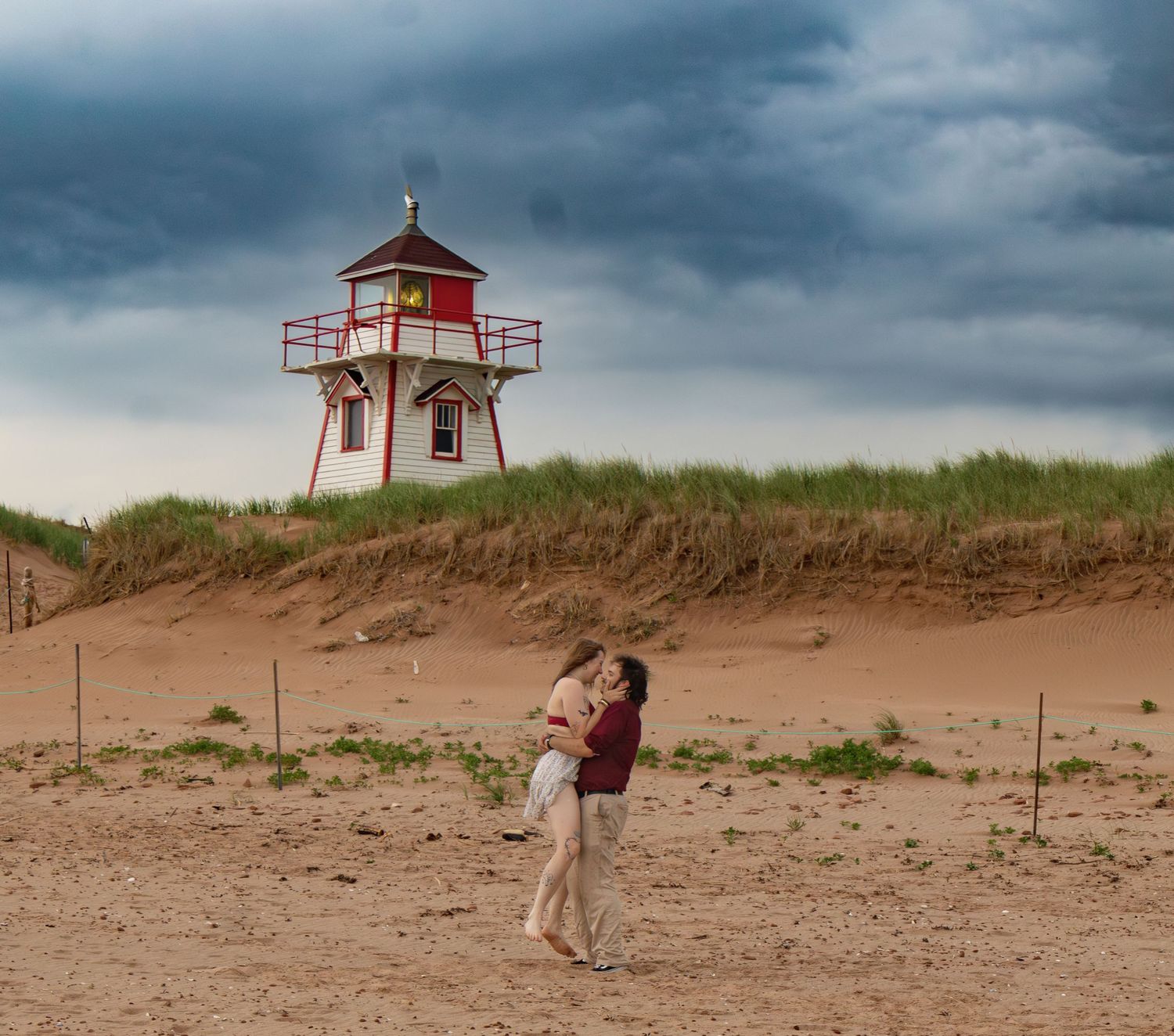 A lighthouse stands on sandy dunes under dramatic stormy clouds.