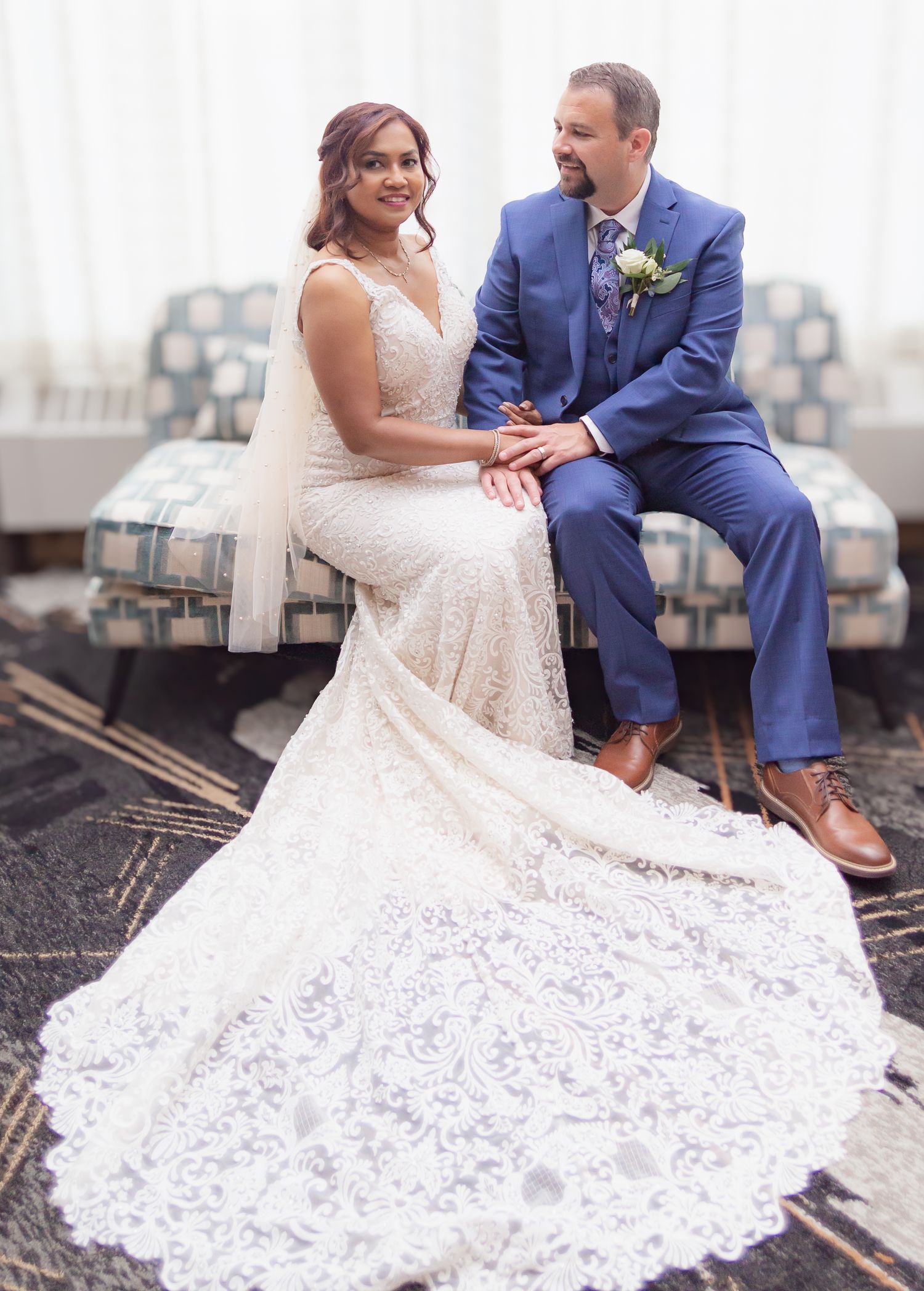 Wedding couple posing with beautiful lace dress spread out on patterned floor.