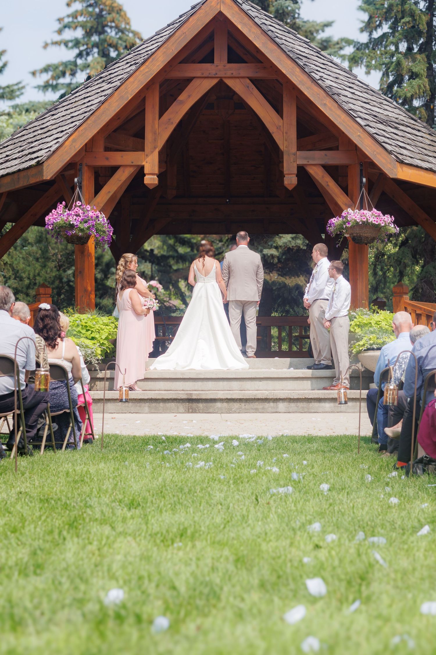 Outdoor wedding ceremony under a wooden pavilion decorated with pink flowers.