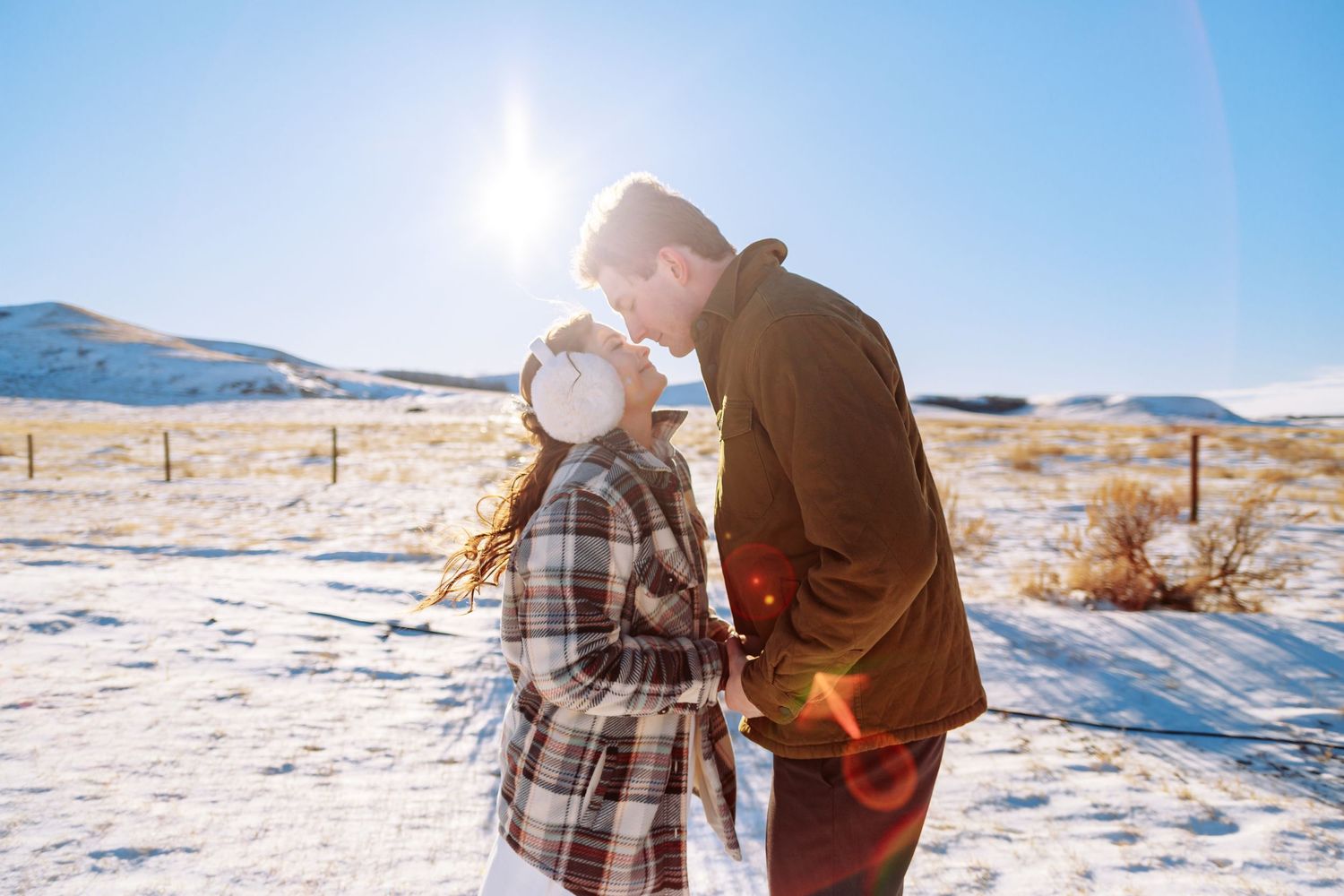 A couple embraces in a snowy winter landscape against a bright blue sky.
