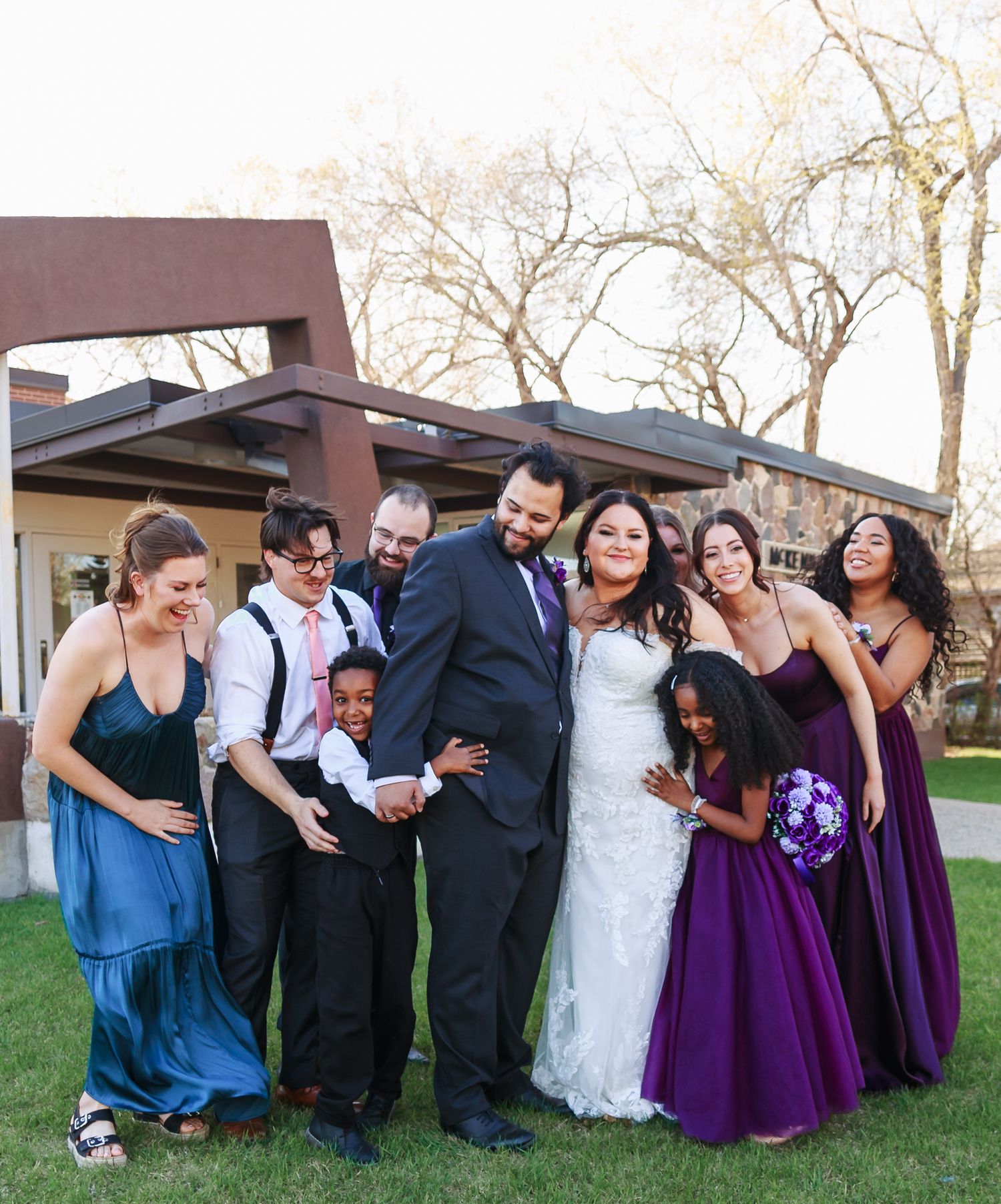 Fun wedding party group photo with bridesmaids in purple dresses on grass.