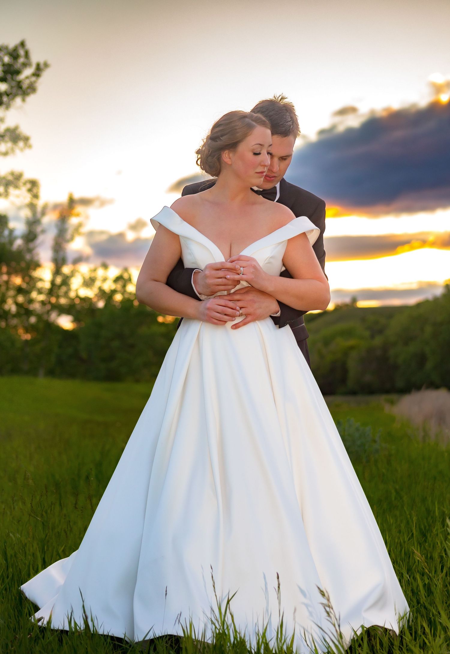 Romantic sunset portrait of newlyweds embracing in a grassy field with flowing white wedding dress.
