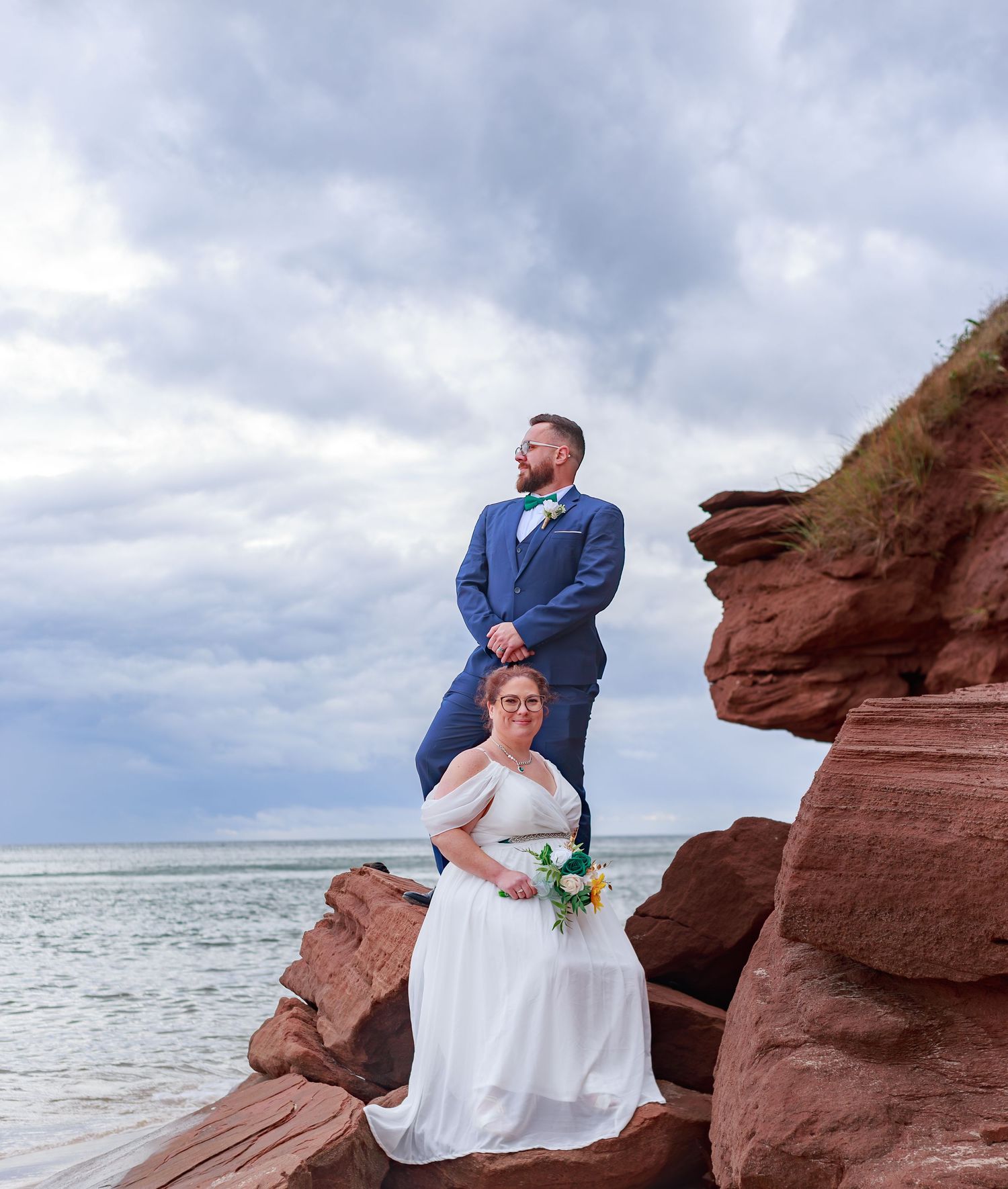 A couple poses on red rock formations by the ocean under cloudy skies.
