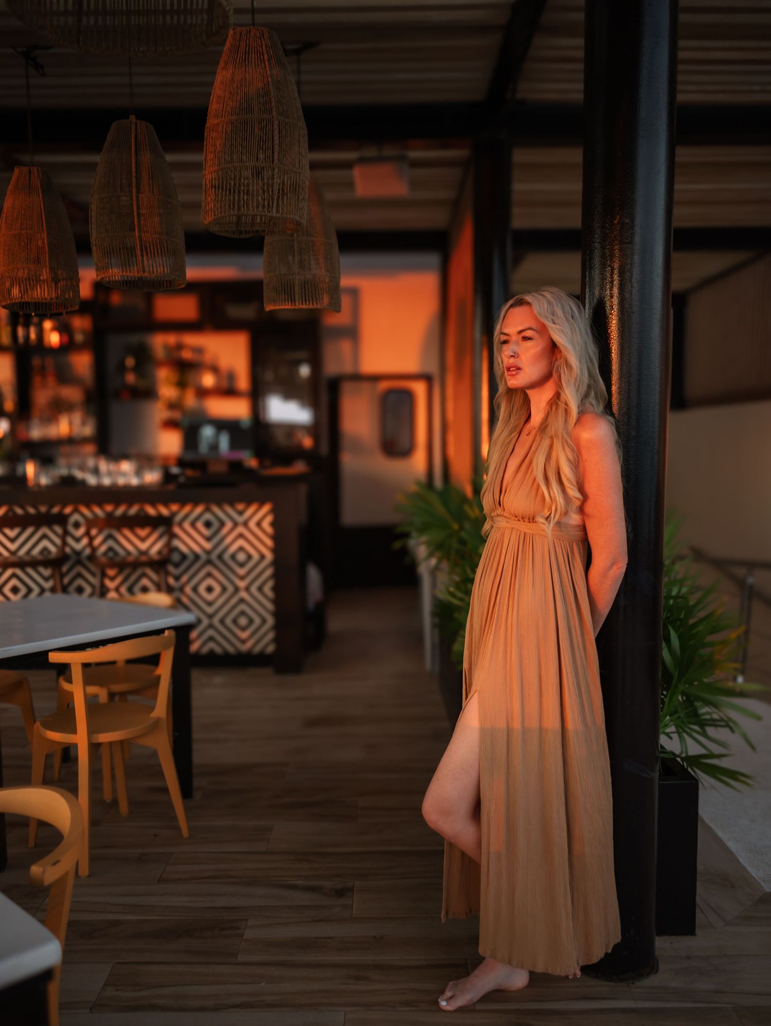 A flowing tan maxi dress catches the warm light in a restaurant with woven pendant lamps and tropical decor.