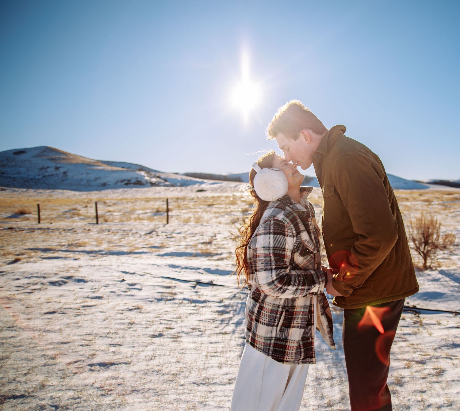 A romantic winter scene with a couple embracing in snow-covered mountains under bright sunlight.