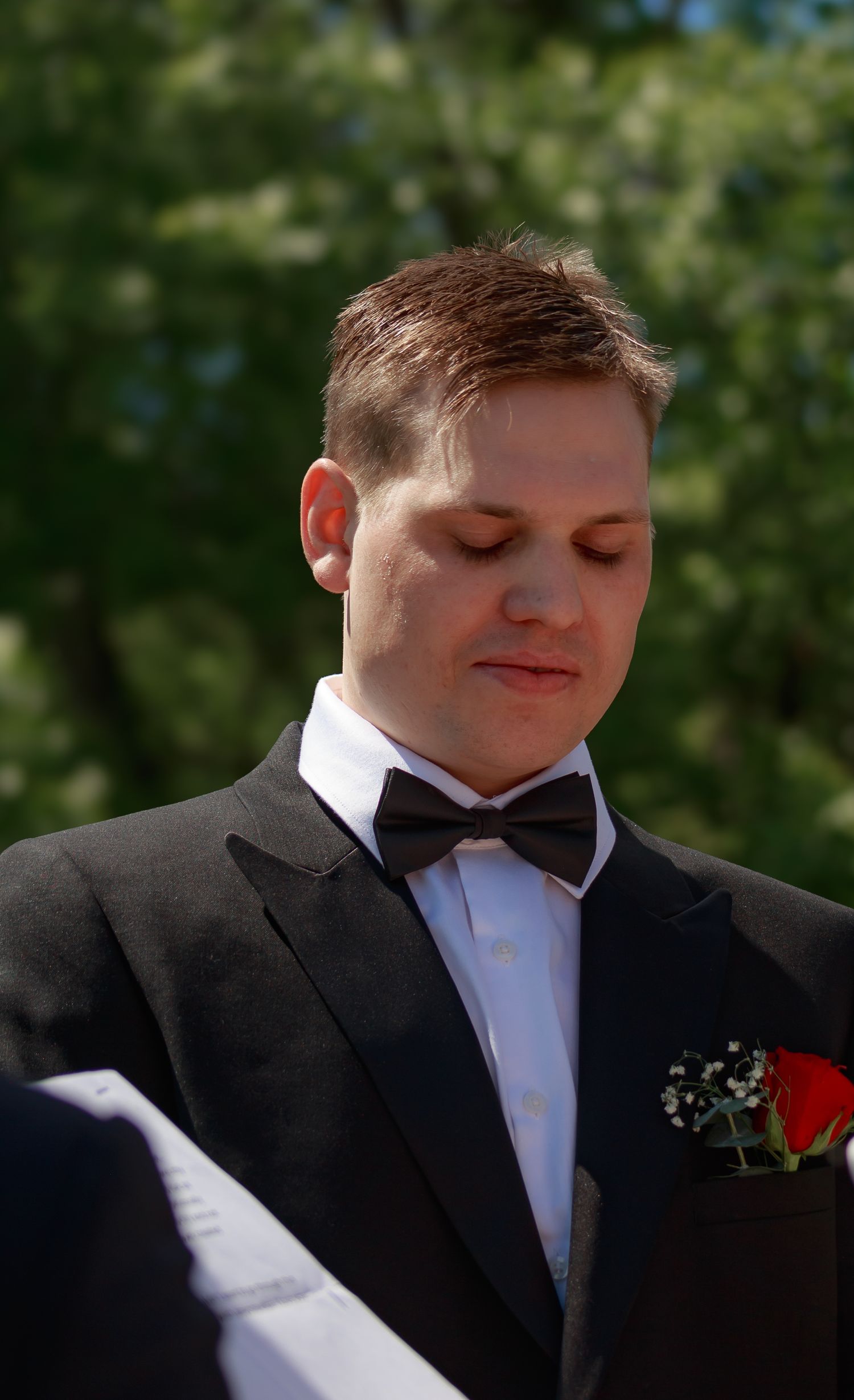 Close-up details of a groom wearing a black tuxedo with red boutonniere at an outdoor wedding.