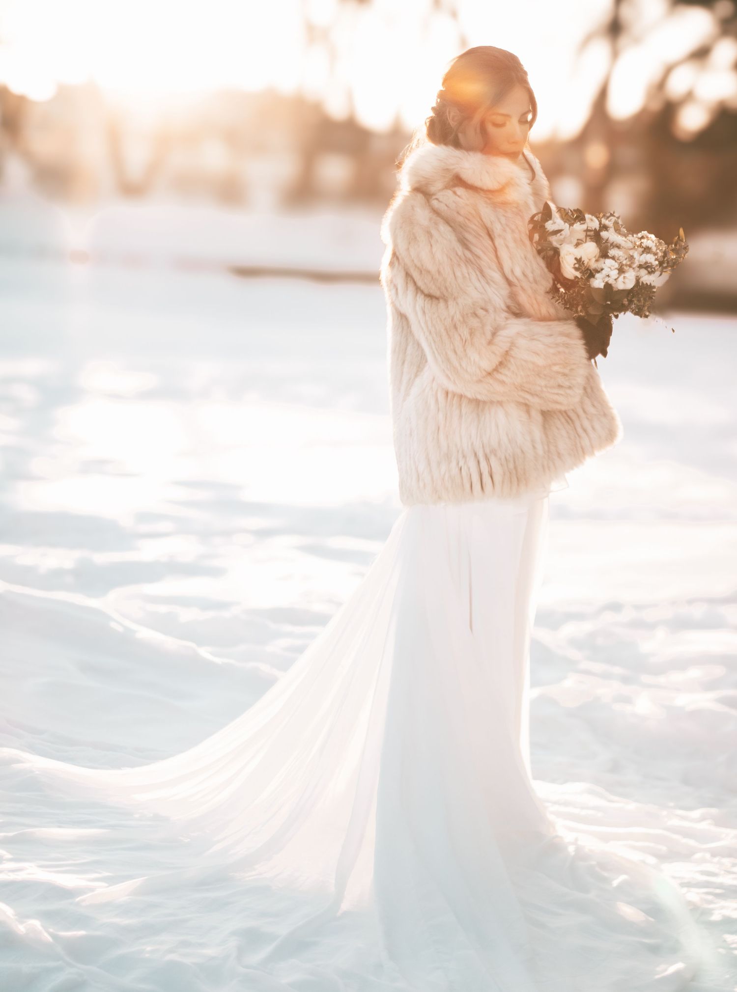 A couple embraces in a snowy winter wonderland setting with golden sunlight filtering through the trees.