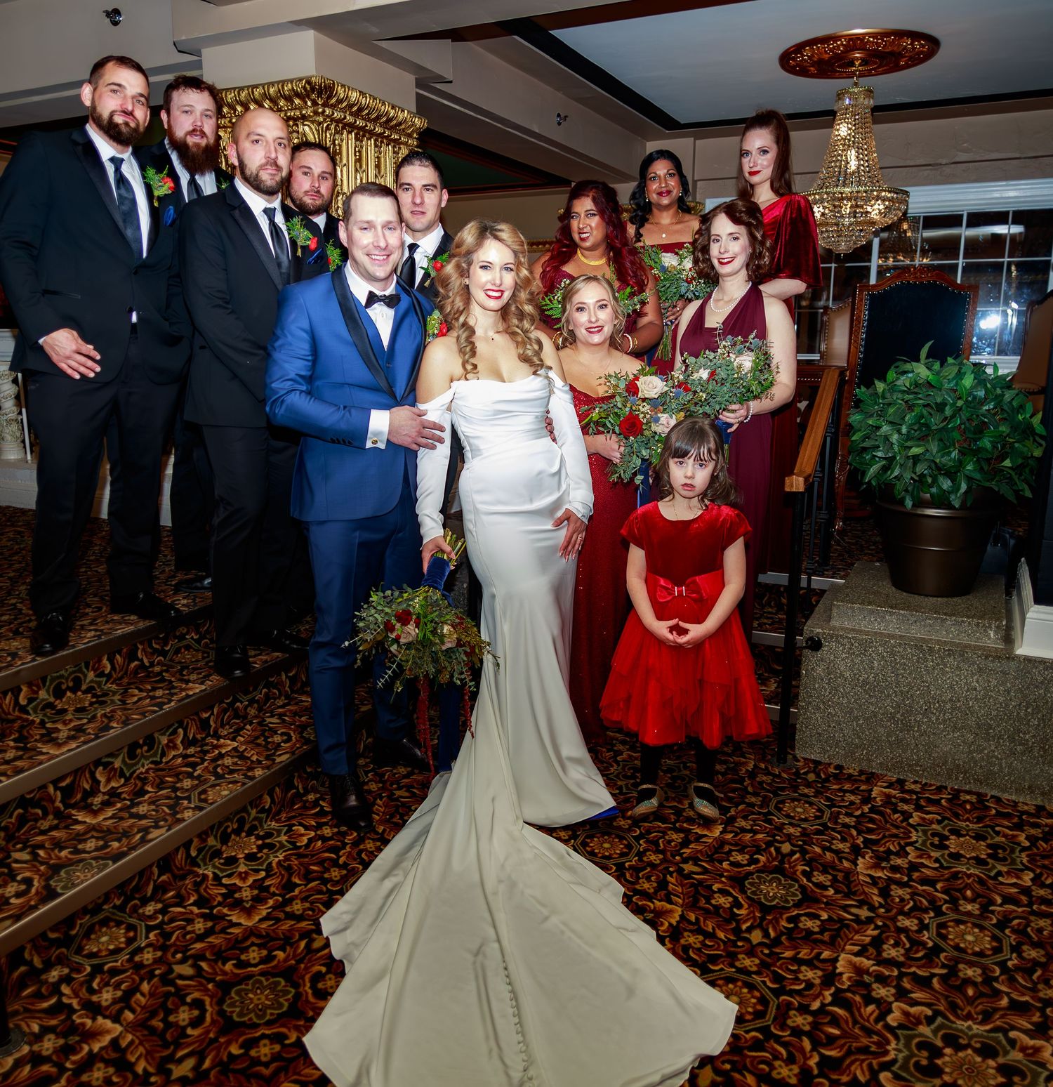 Wedding party poses for formal photos in an elegant venue with burgundy carpet and ornate decor.