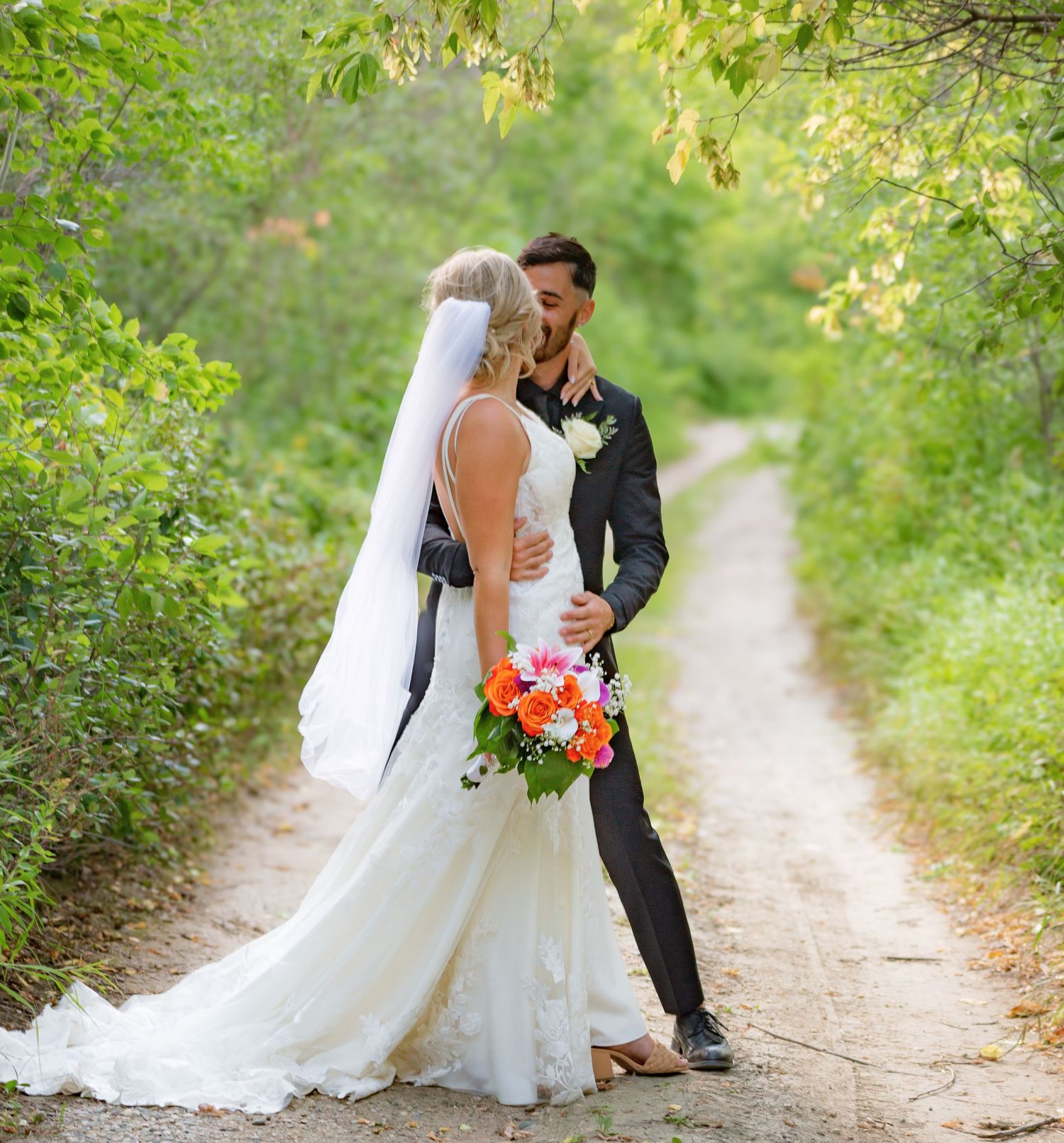 Romantic wedding portraits on a sunlit nature trail surrounded by green foliage.
