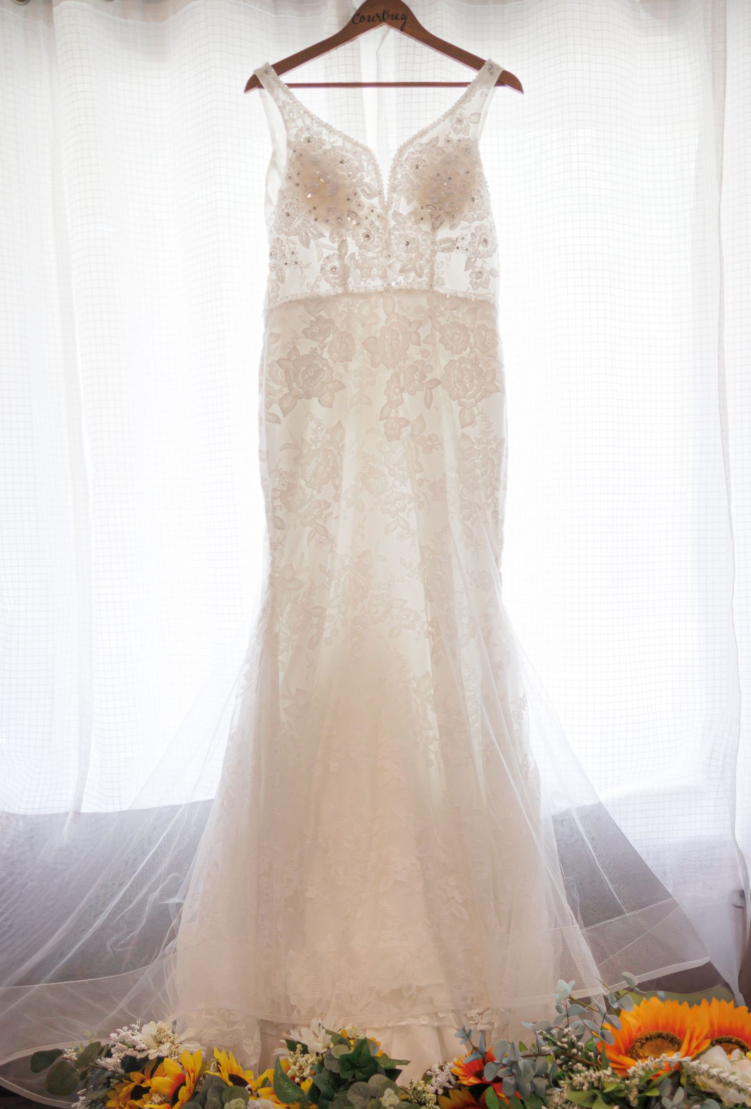 Elegant lace wedding dress hanging against white backdrop with sunflowers at base.