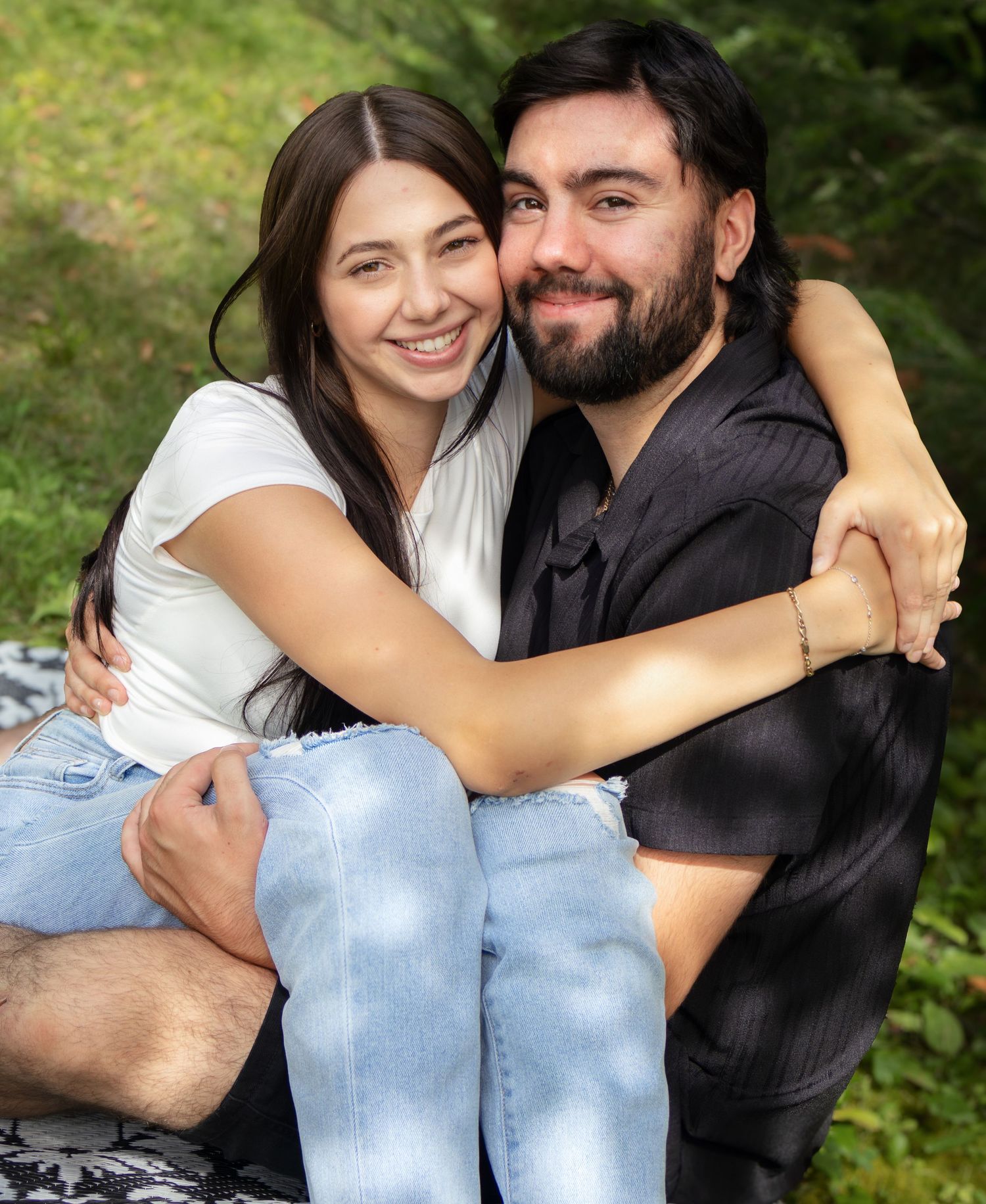 A couple in casual attire embraces while sitting outdoors in a natural setting.