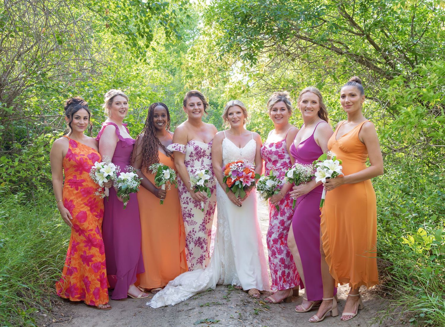 Wedding party in colorful orange and purple dresses posing outdoors against lush green foliage.