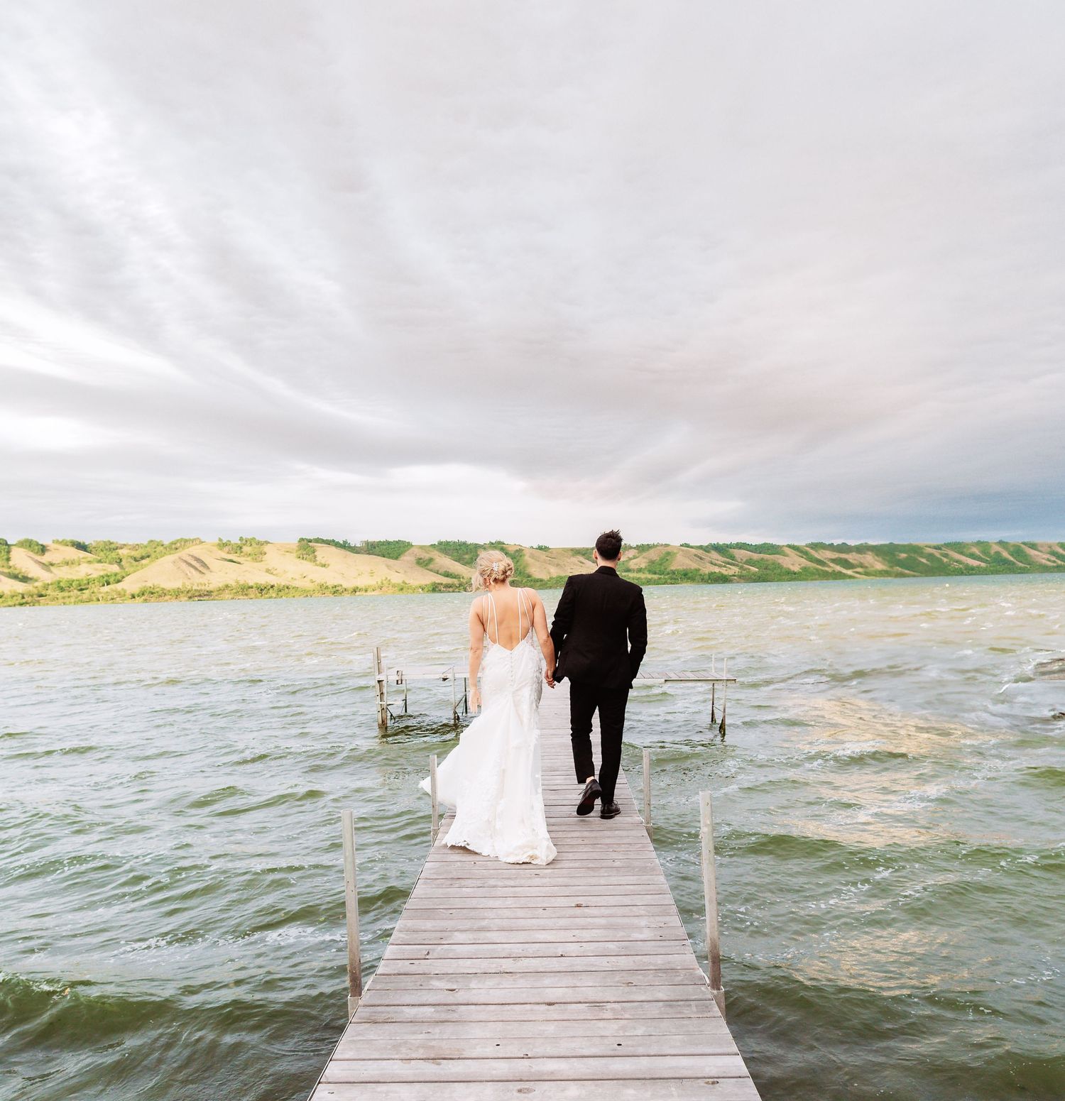 Romantic couple walking on a wooden dock overlooking a serene lake on their wedding day.