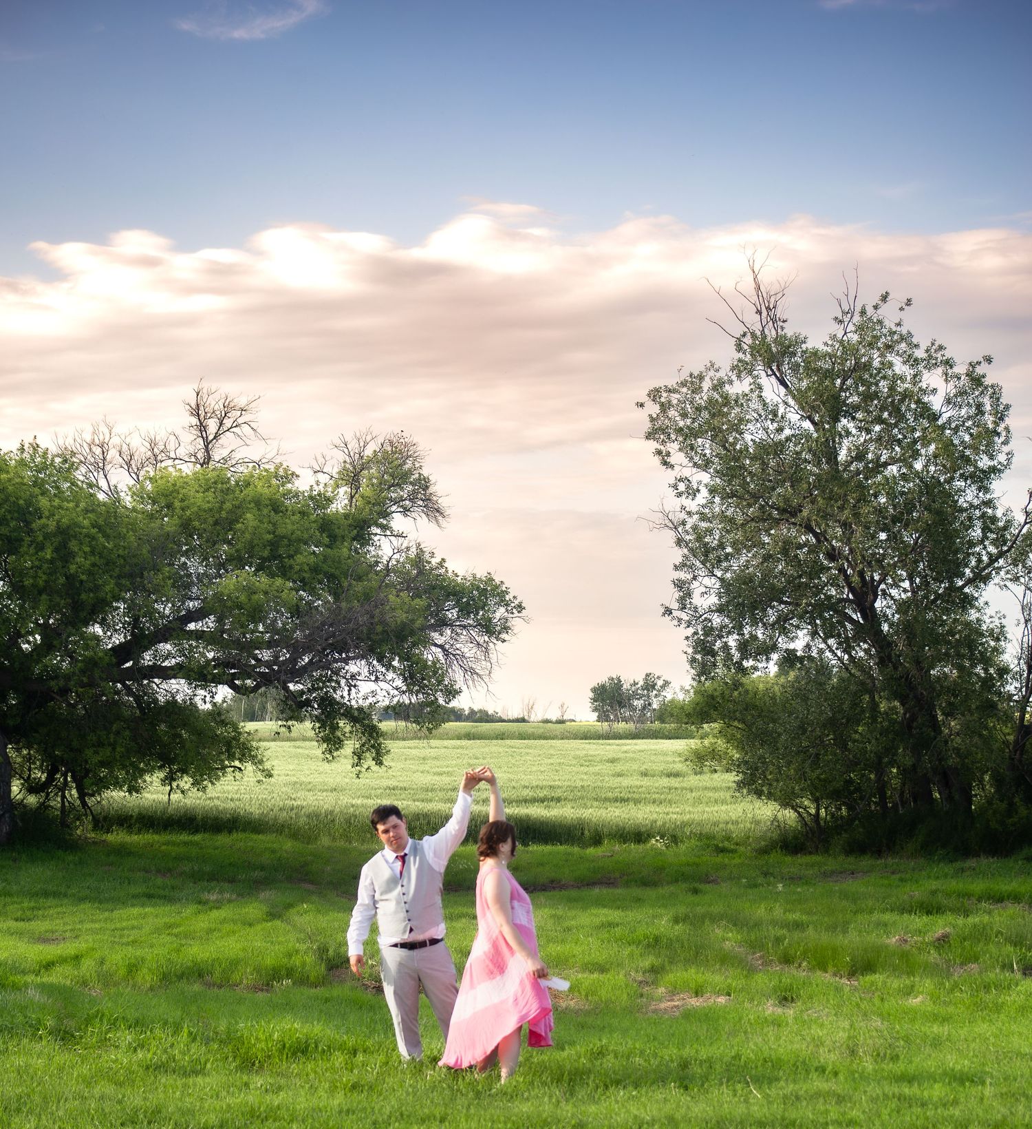 A couple dances together in a dreamy open field with trees and sunset skies in the background.